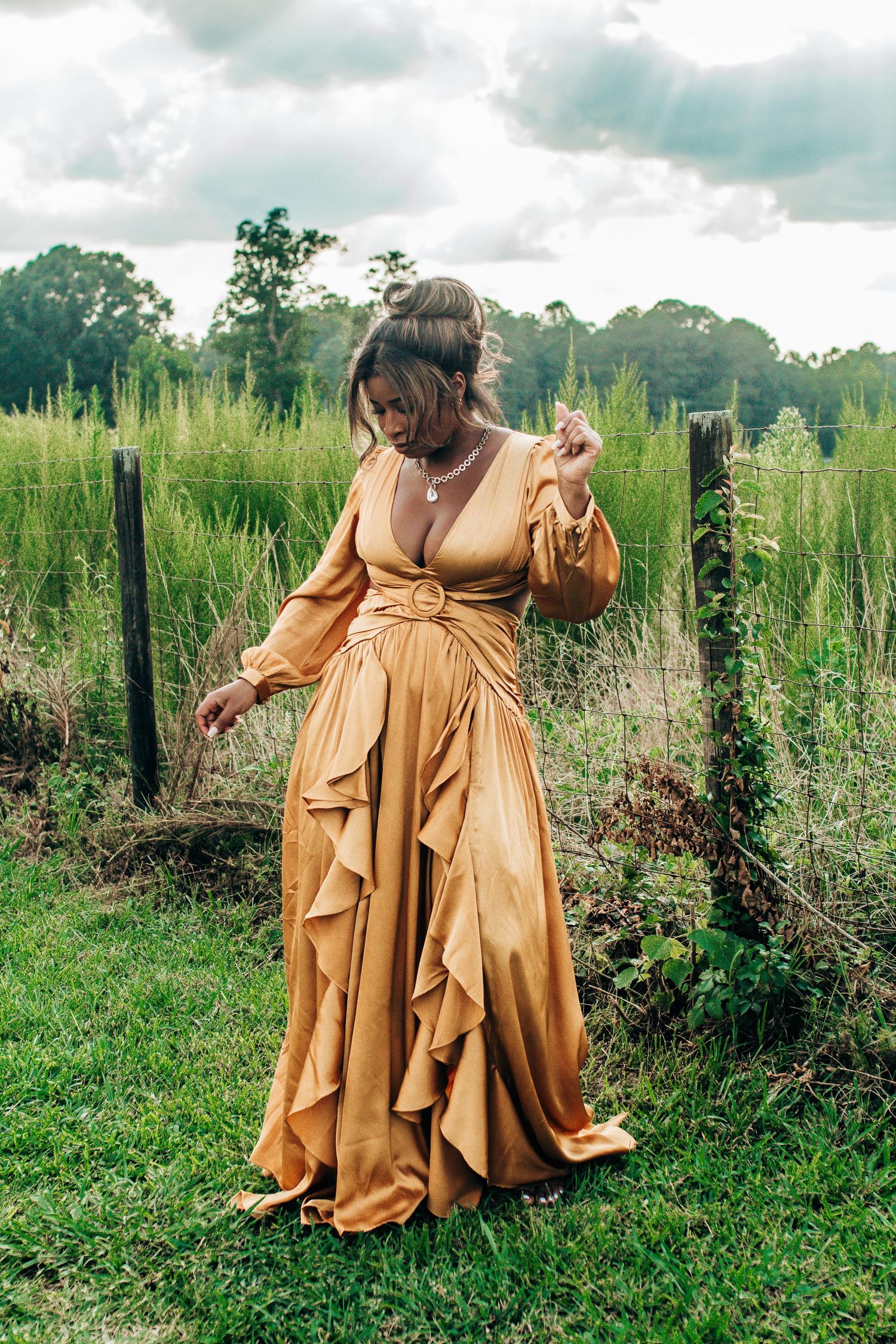 A person in a flowing caramel-colored dress twirls gracefully in a lush green meadow on a cloudy day.