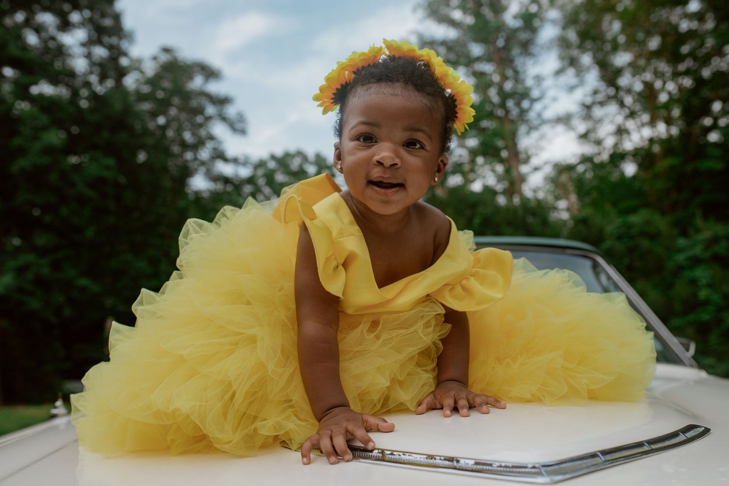 A child in a bright yellow tulle dress and matching floral crown poses gracefully on a porch swing outdoors.