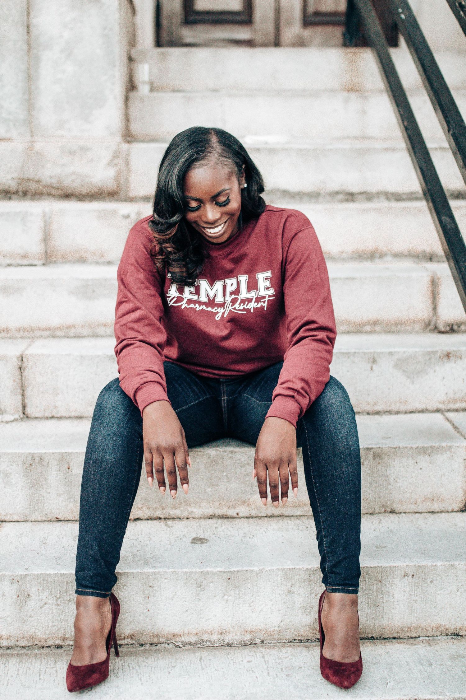 Person wearing maroon college sweatshirt and jeans sits casually on concrete steps in urban setting.