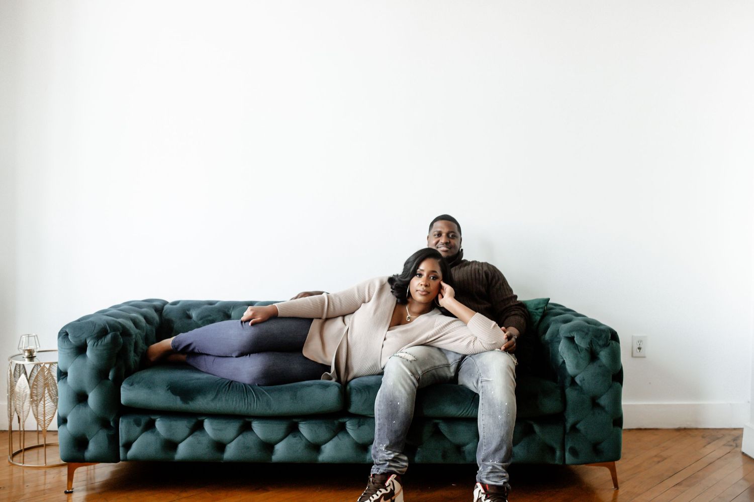 Wide shot of a couple lounging on a tufted teal sofa in a bright room.