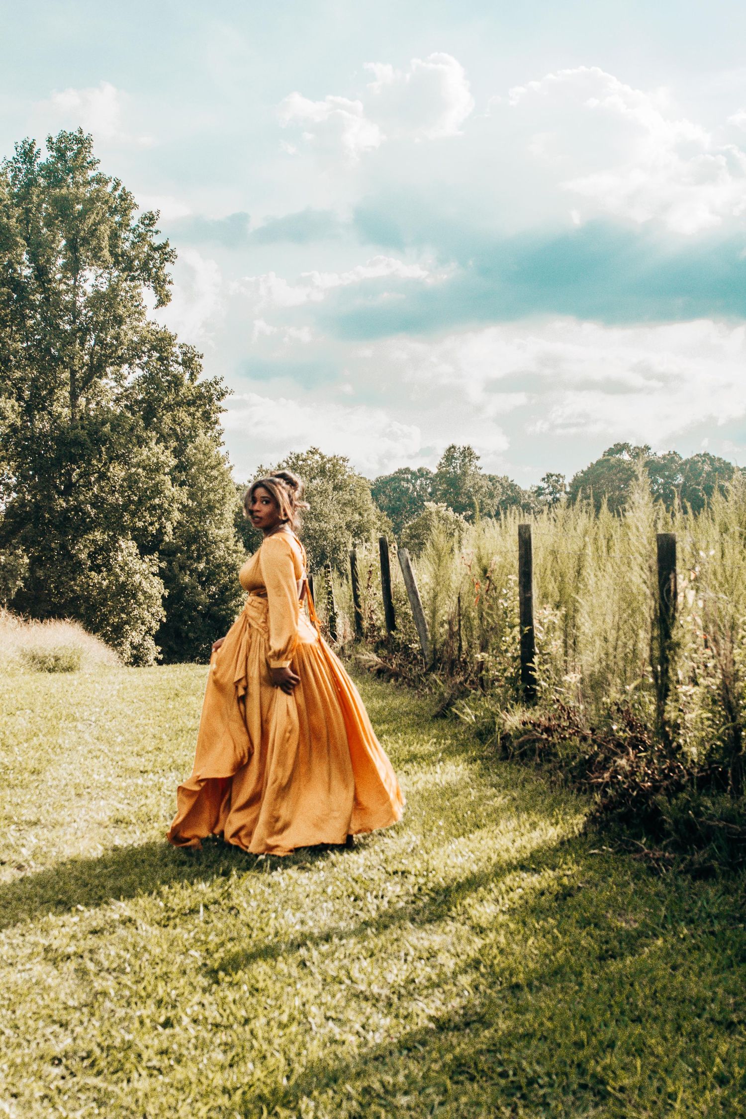 A person in a flowing mustard yellow dress walks along a sunlit garden path bordered by trees and fence posts.
