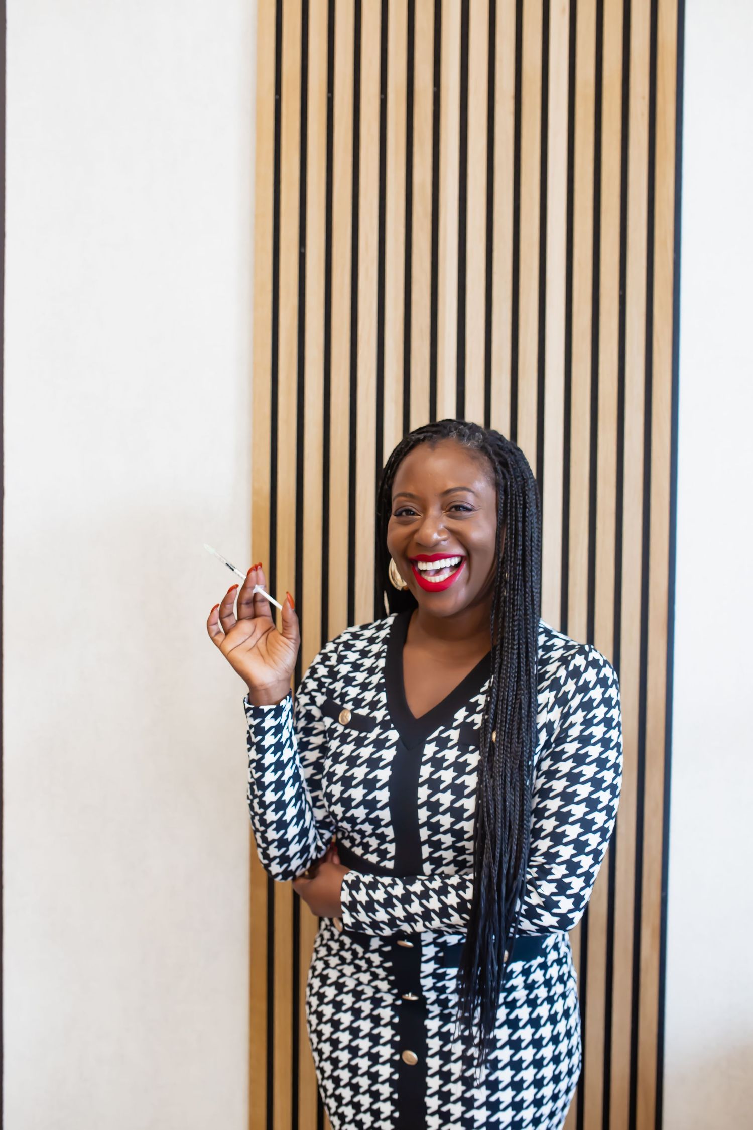 A person in a black and white houndstooth dress stands smiling against a wooden slat wall.