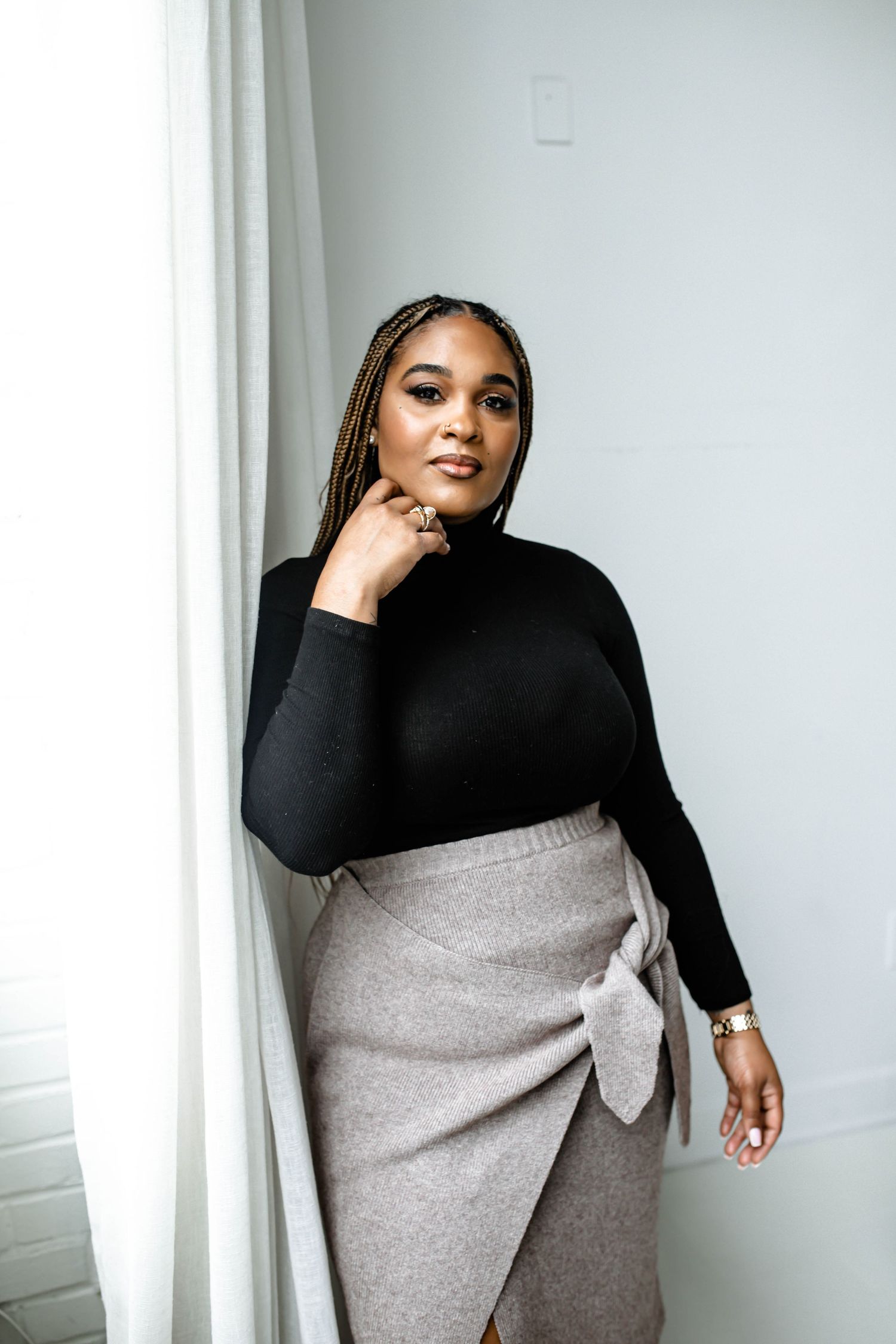Stylish portrait against white wall wearing black turtleneck and gray wrap skirt in modern minimalist setting.