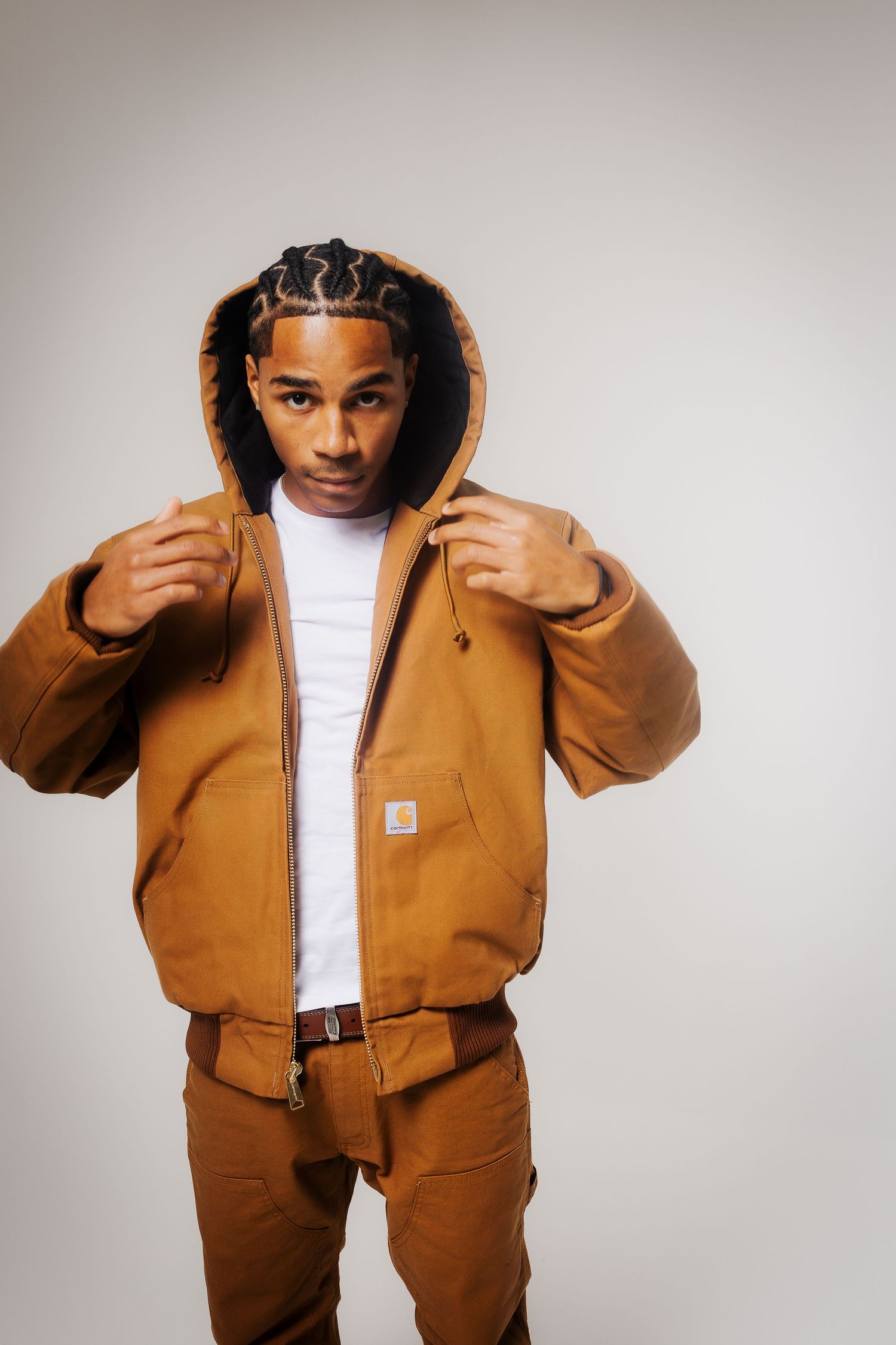 A person wearing a brown Carhartt workwear jacket and matching pants poses against a light gray background.