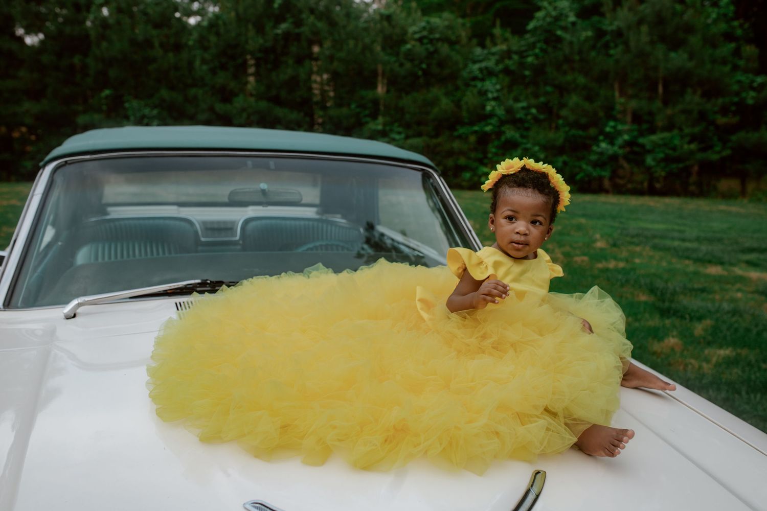 A small child in a bright yellow tulle dress poses on the hood of a classic white car in a grassy outdoor setting.