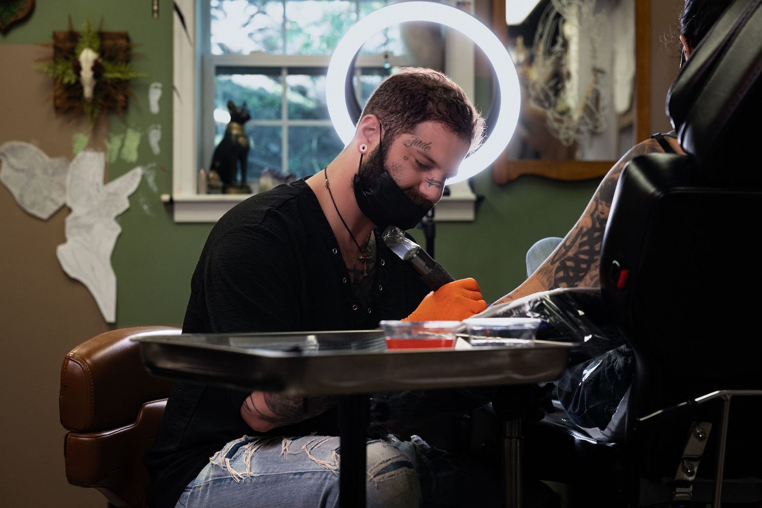Bearded tattoo artist working on a client in a decorated tattoo studio, wearing orange gloves under ring light.