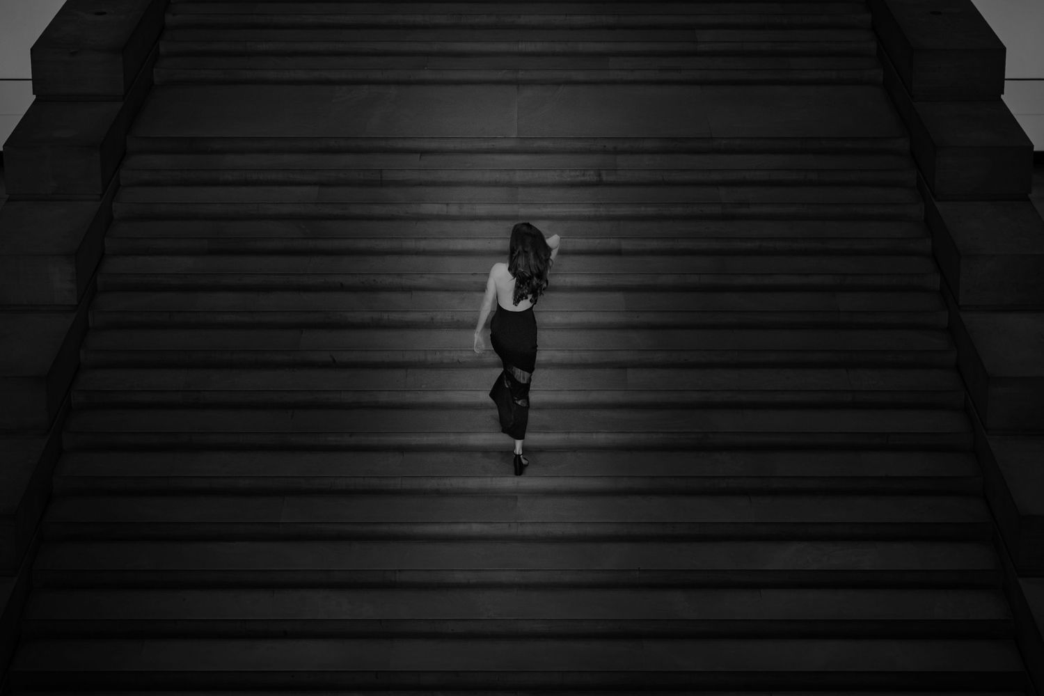 Black and white photo of a woman in black outfit ascending wide stone stairs, viewed from above.