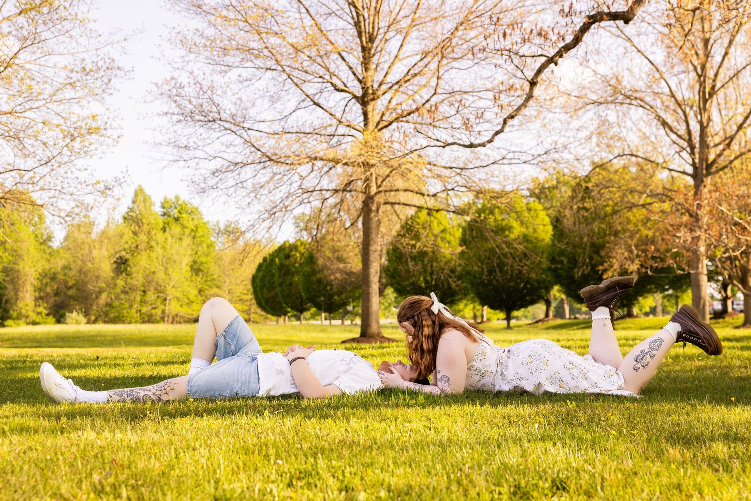 Couple lying on green grass in a sunny park surrounded by trees, enjoying a romantic outdoor moment together.