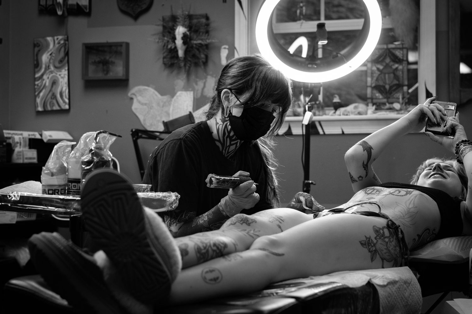 Black and white photo of a tattoo artist working on a client in a professional tattoo studio with ring light.