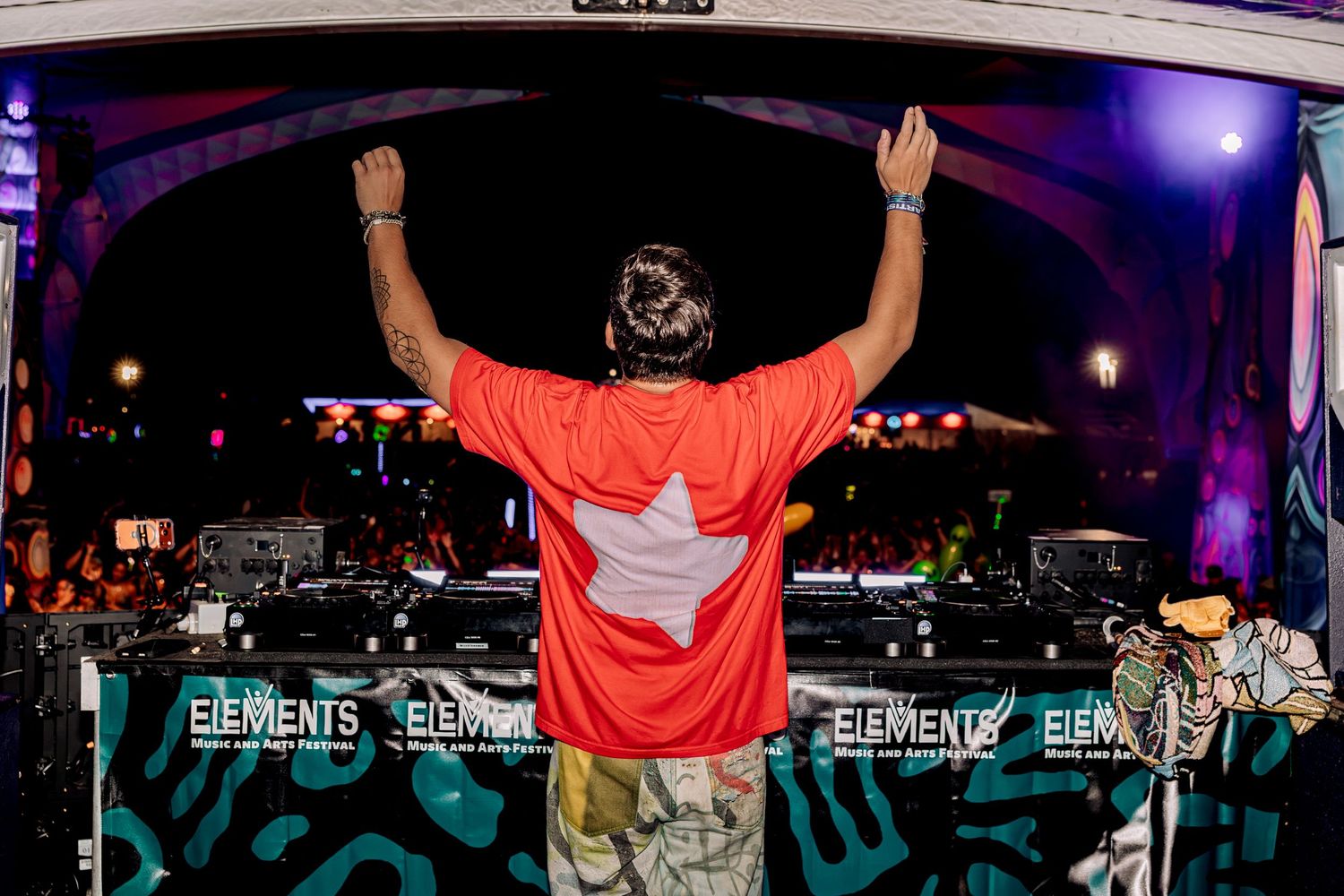 DJ performing on stage at Elements festival, arms raised in red shirt behind turntables at night.