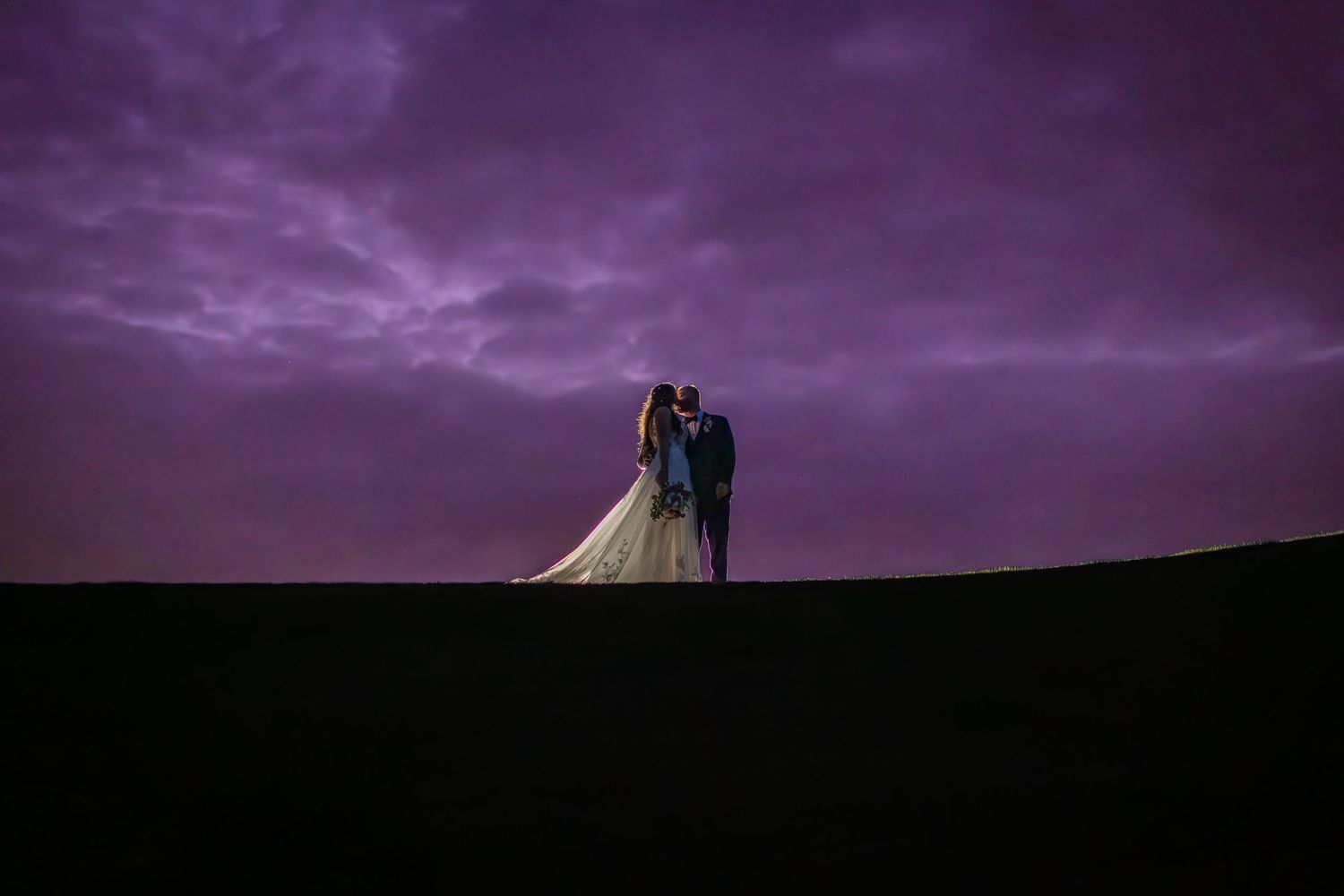Bride and groom silhouetted against a dramatic purple twilight sky on their wedding day.