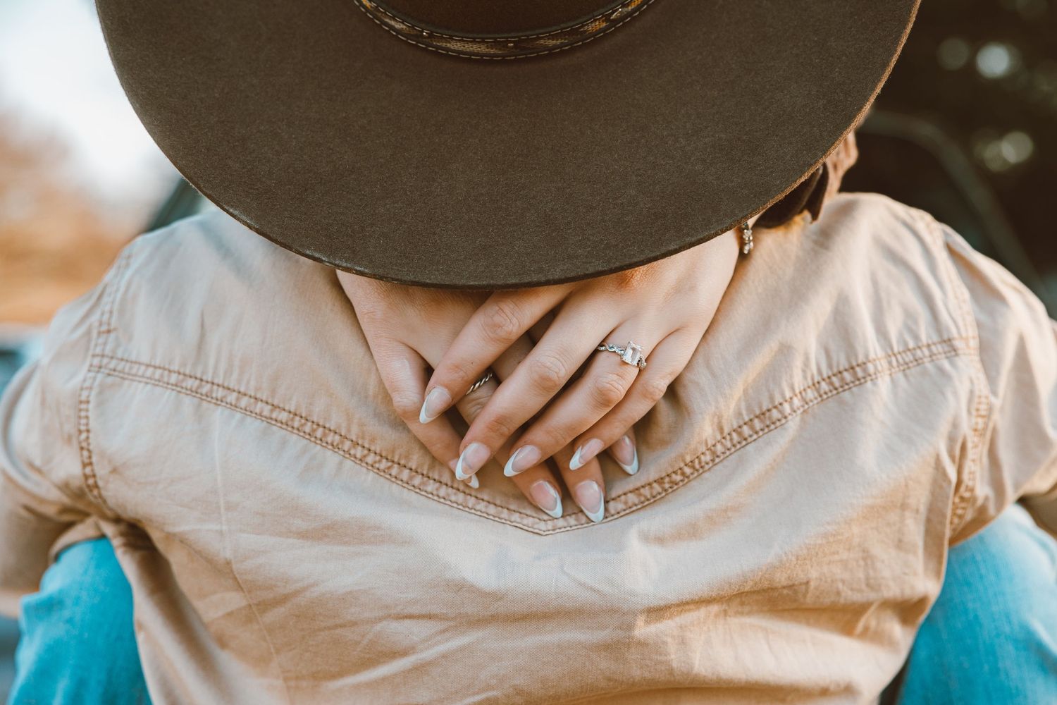 Person wearing a wide-brim hat and beige shirt, showing off a diamond engagement ring with manicured nails.