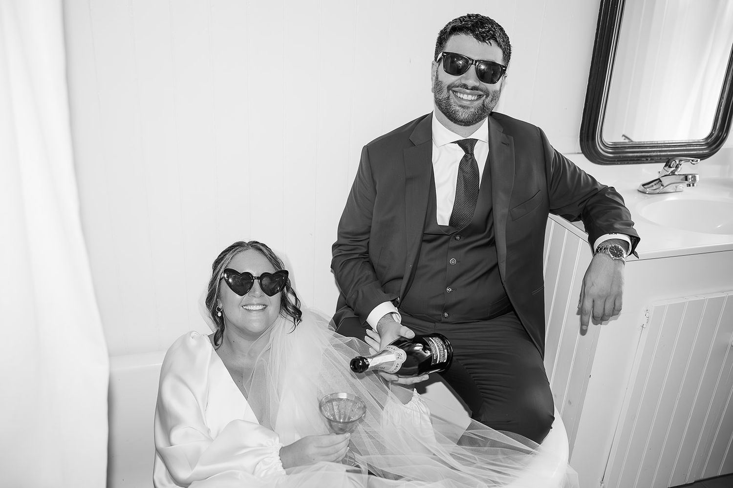 Black and white photo of a smiling bride and groom in sunglasses posing together on their wedding day.