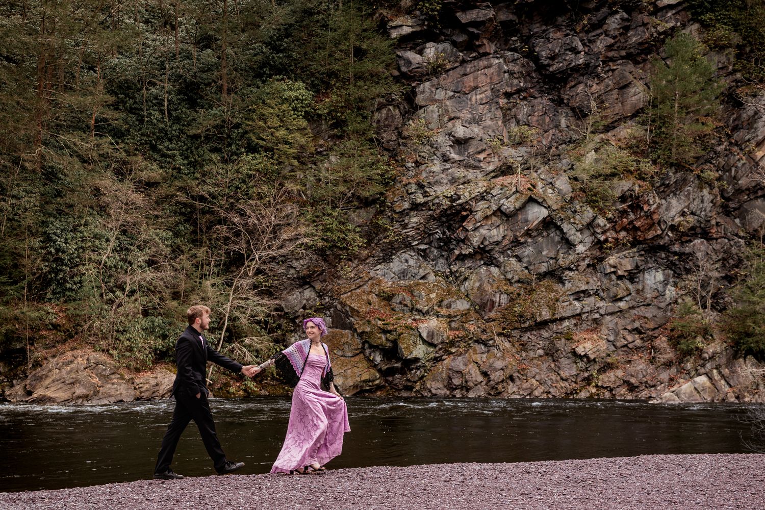 Couple in formal wear walks hand-in-hand along a rocky riverbank with a forested cliff backdrop.