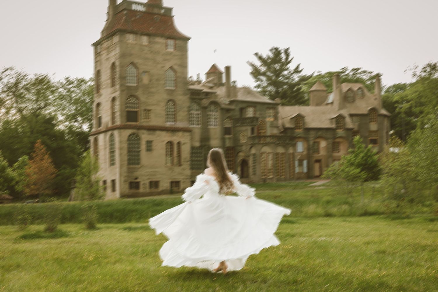 Woman in flowing white dress twirling on green lawn before a grand historic stone castle.