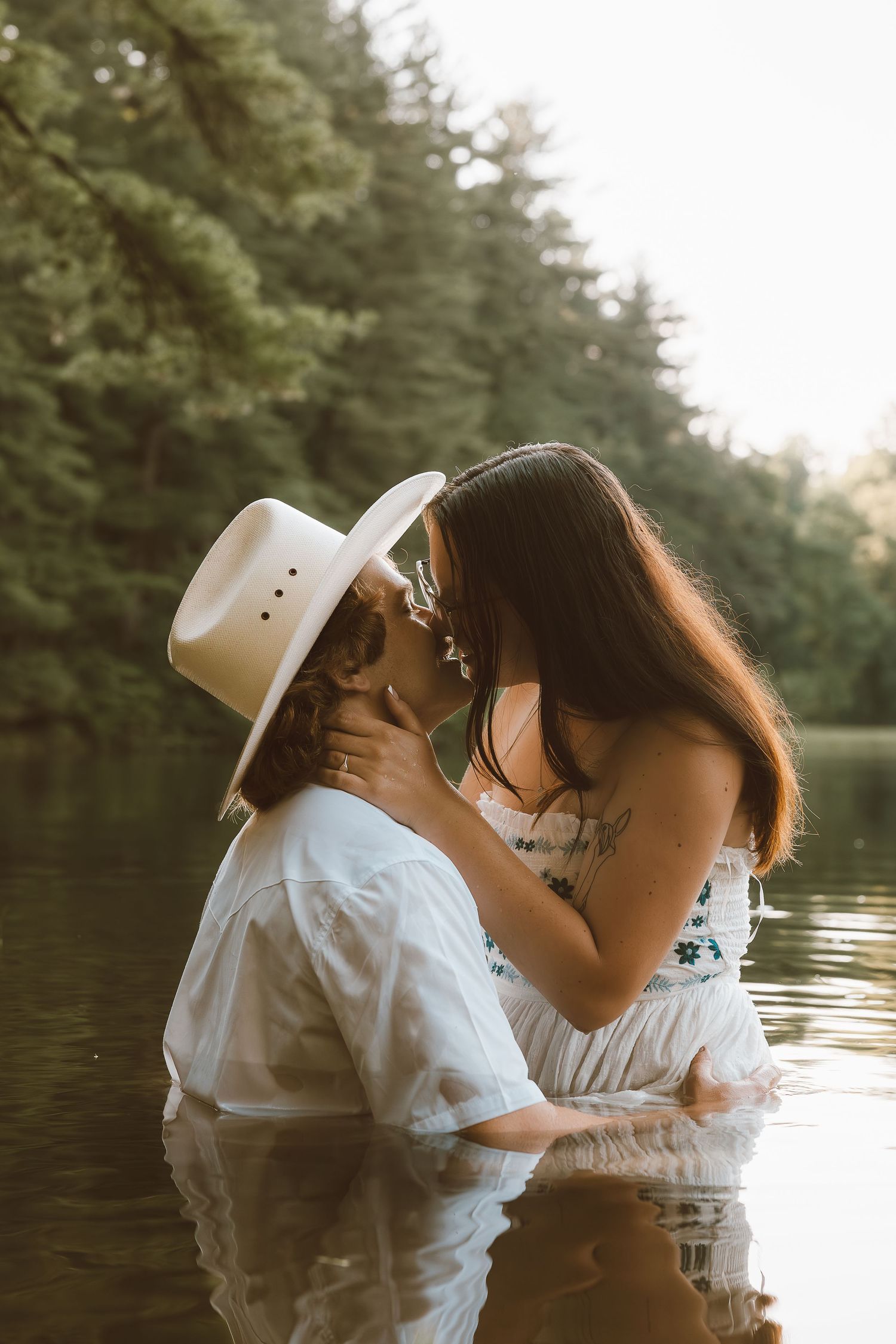 Romantic couple sharing intimate moment in white clothing by a lake at sunset.