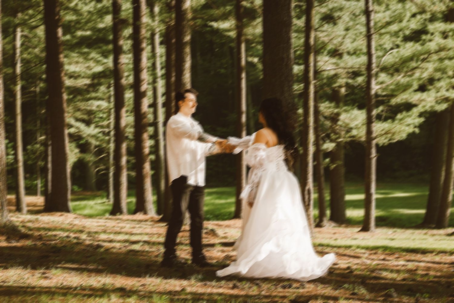 A bride and groom hold hands in a sunlit forest clearing, the bride wearing an elegant off-shoulder white gown.