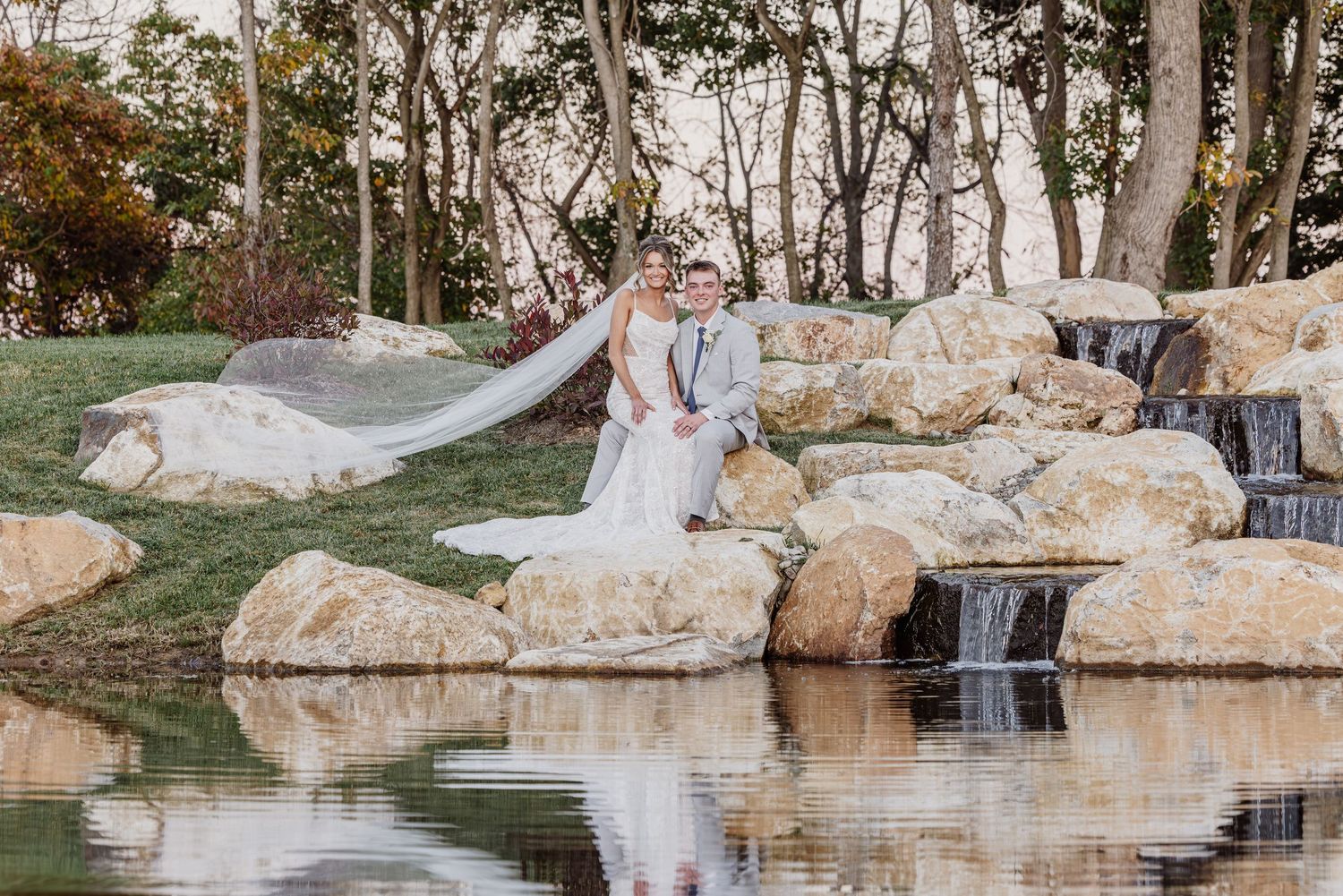 Bride and groom pose on rocky waterfall backdrop in lush garden setting for elegant outdoor wedding portrait.