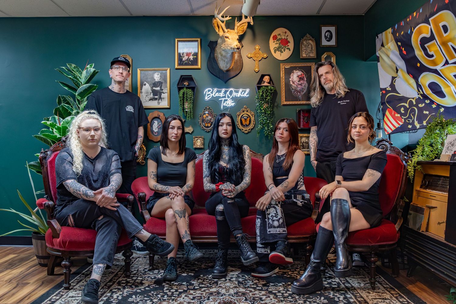 A group sits on a red couch in a tattoo parlor with artwork and decorations visible on the walls.