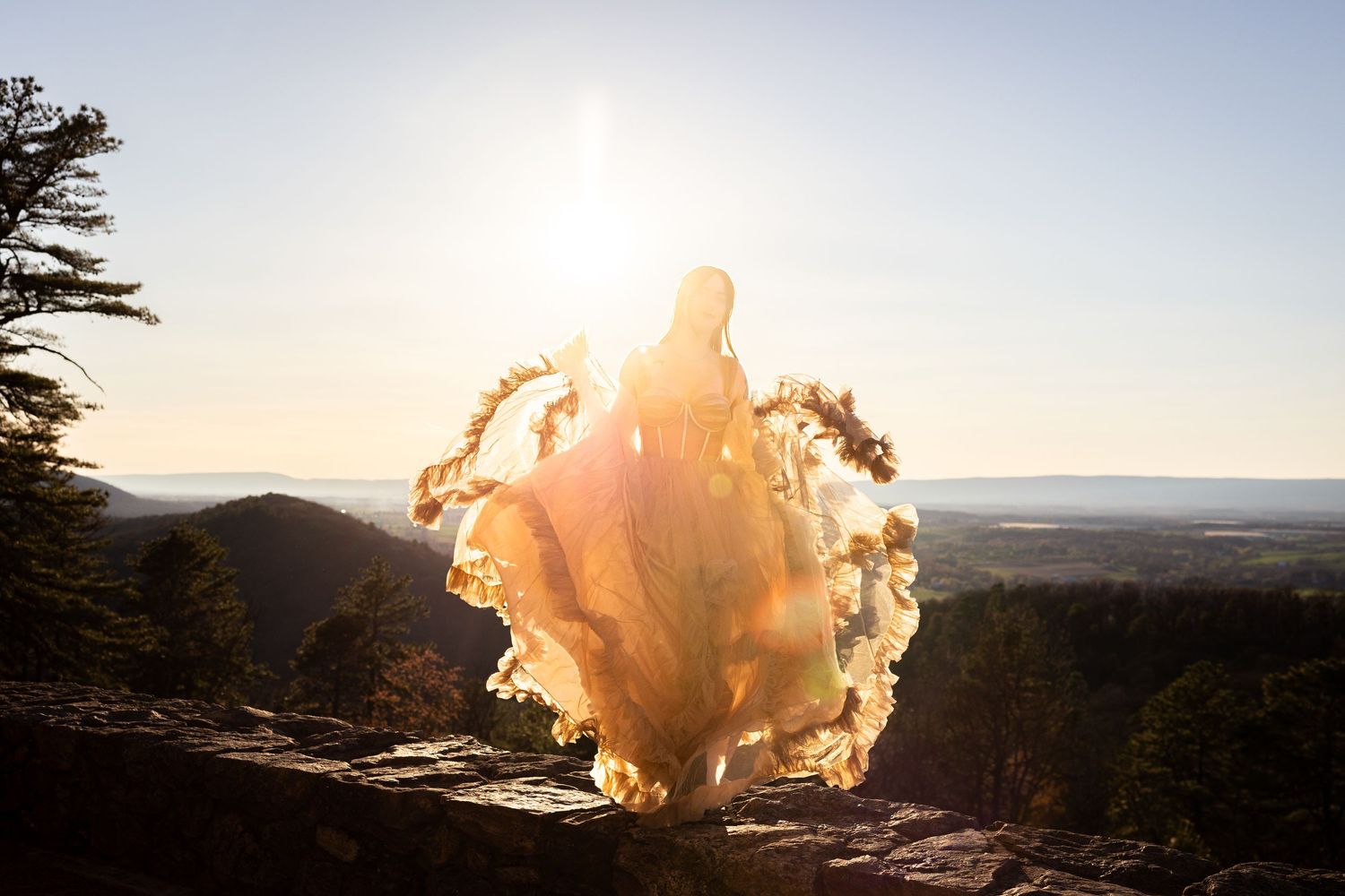 A bride in a flowing golden gown twirls on a rocky overlook at sunset with scenic valley views behind her.
