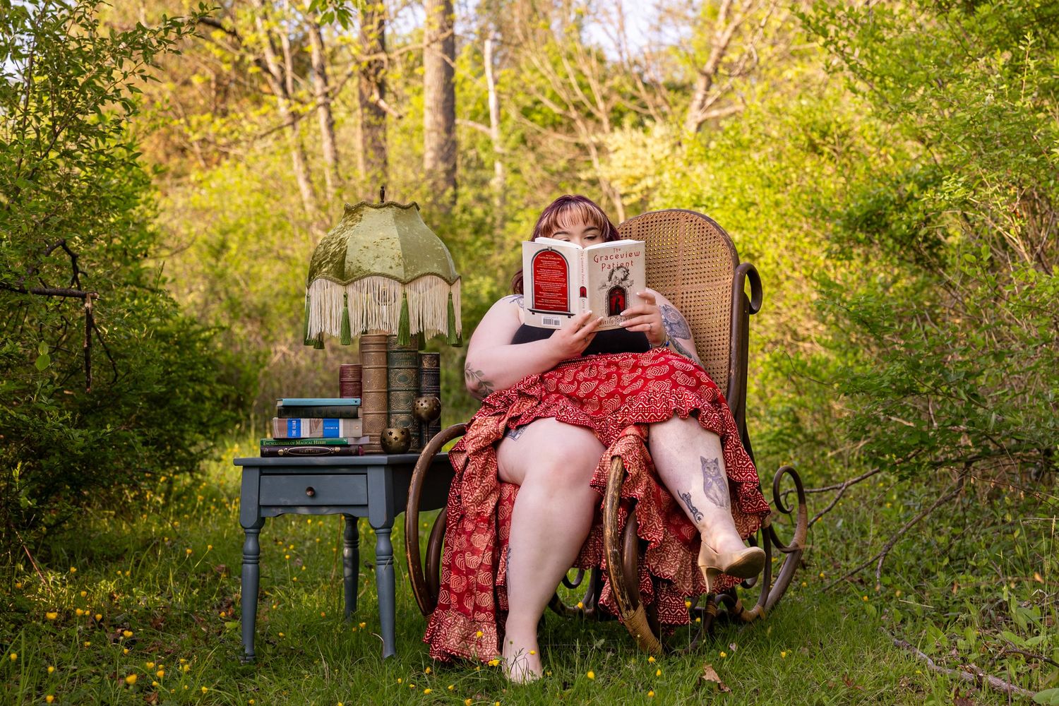 Woman in red dress reading a book in vintage wicker chair outdoors, surrounded by lush green trees and a blue side table.