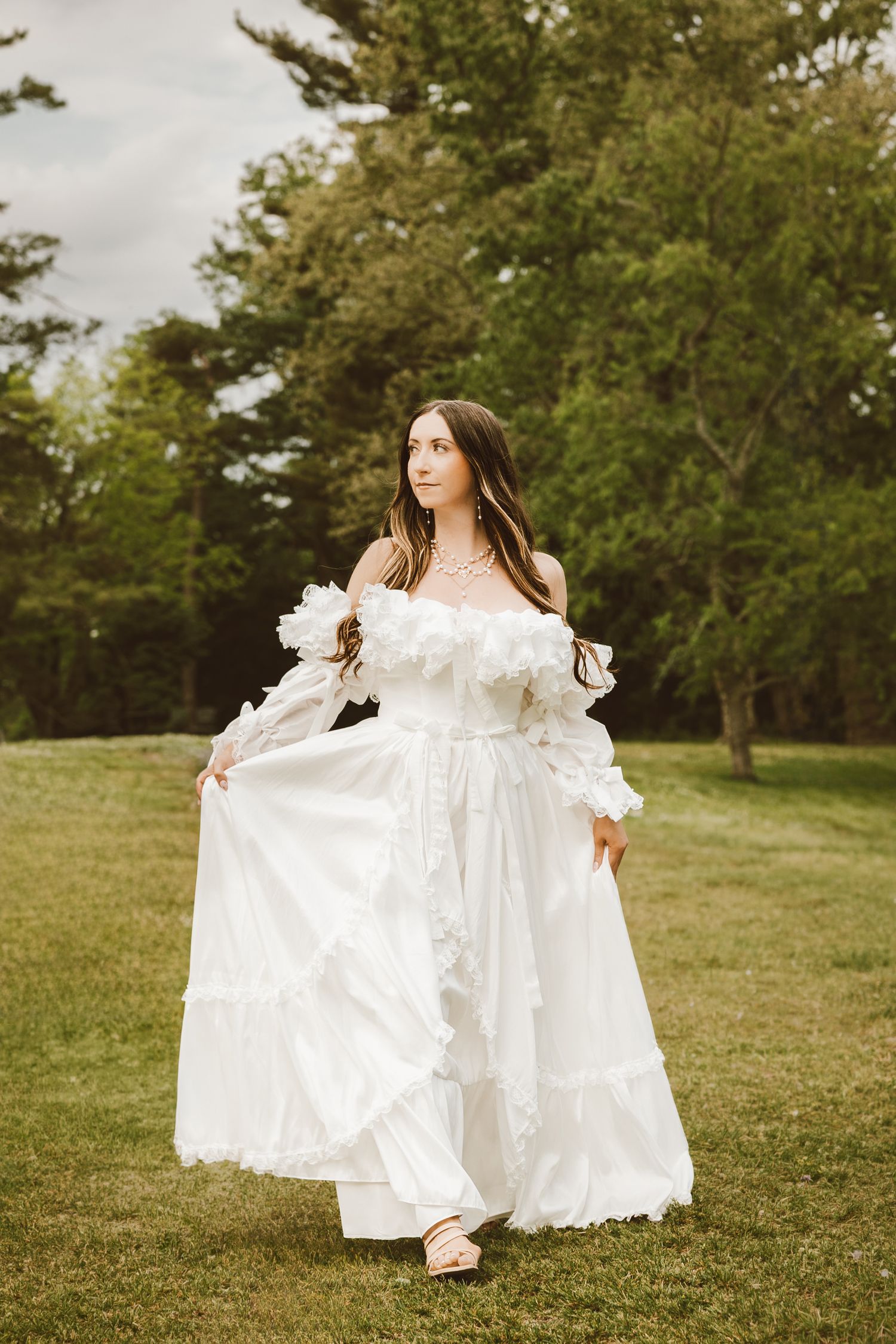 Woman in white ruffled off-shoulder gown posing gracefully on green lawn with trees in background.