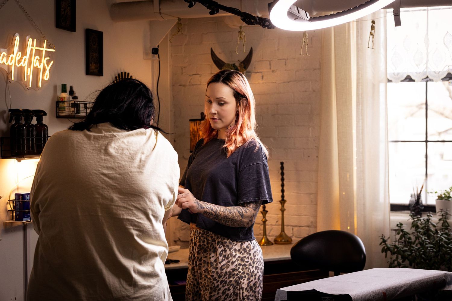 Two women in a cozy tattoo studio, one with devil horns headband and leopard skirt getting assistance from a tattoo artist.