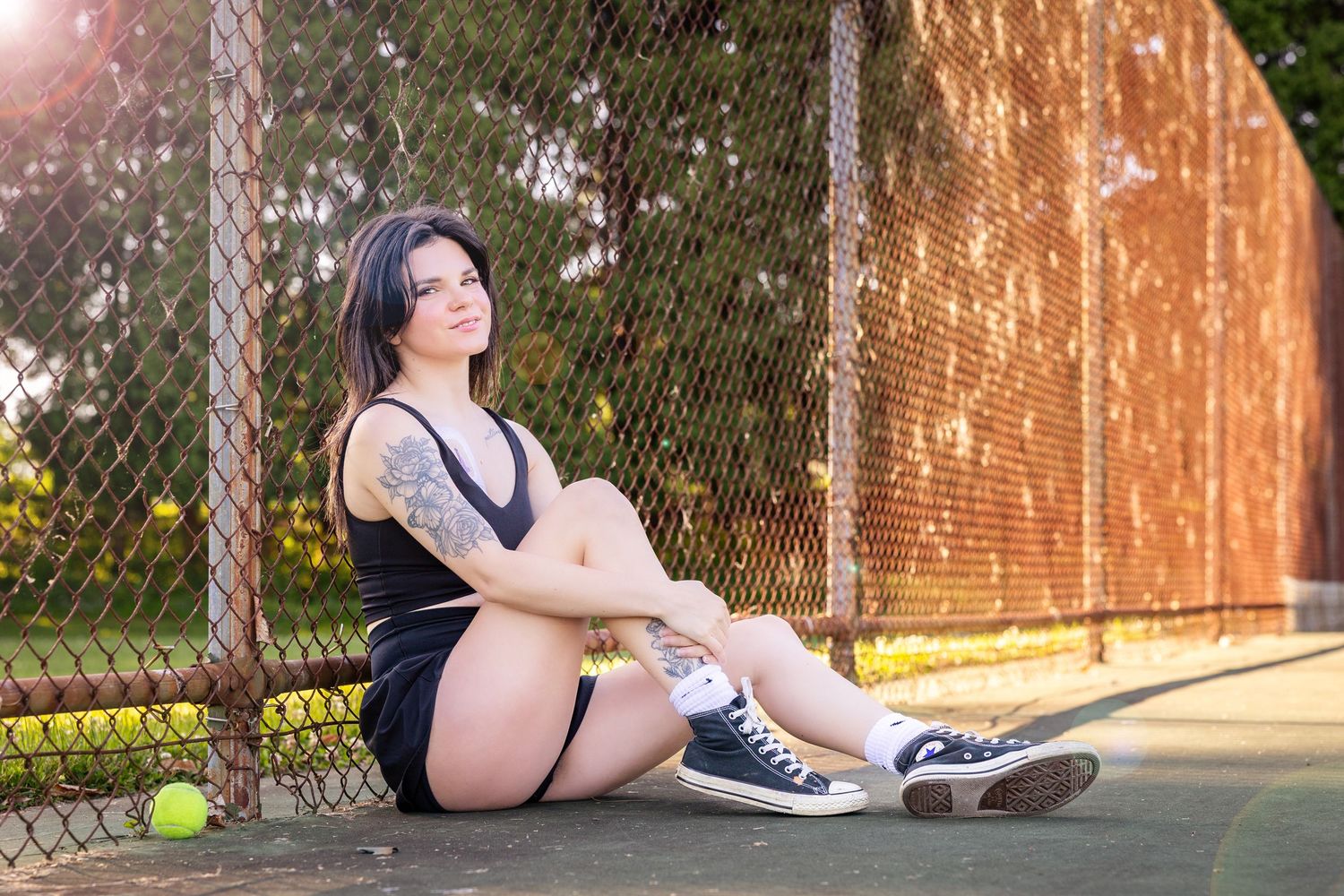 Woman with tattoos sitting on tennis court in black outfit and sneakers, leaning against chain-link fence outdoors.