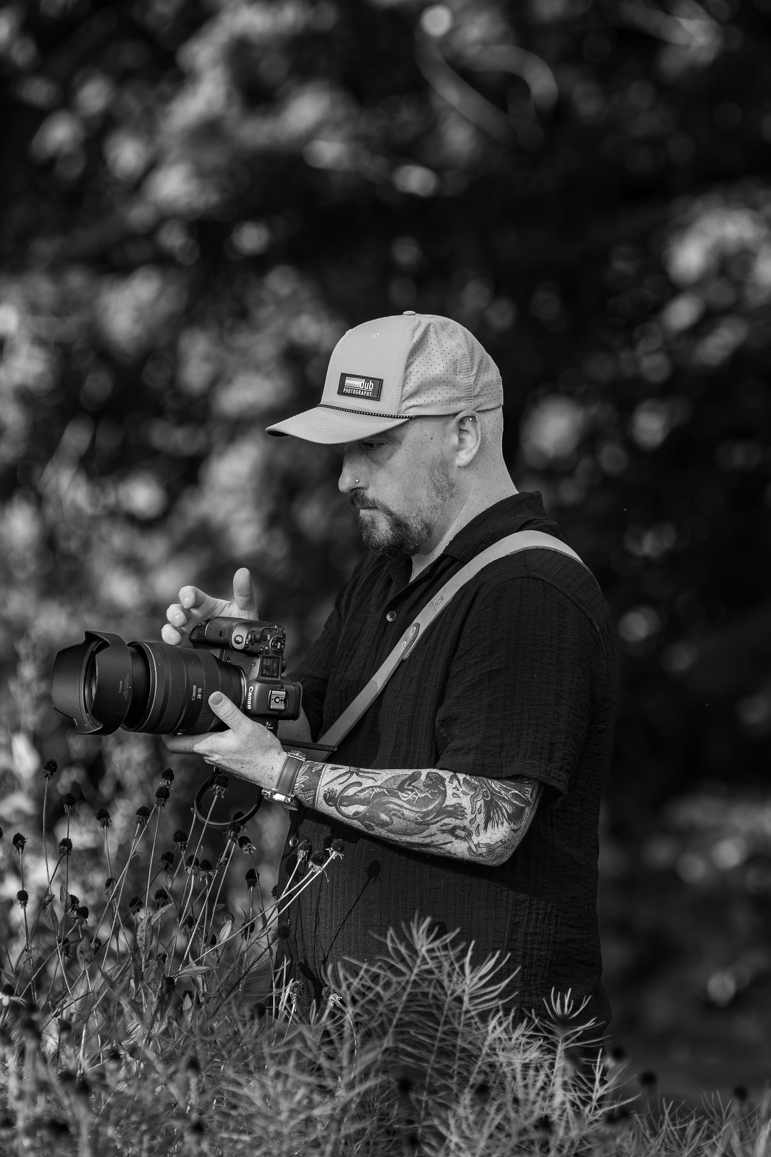 Black and white photo of photographer with tattoos and baseball cap using professional camera in nature setting.