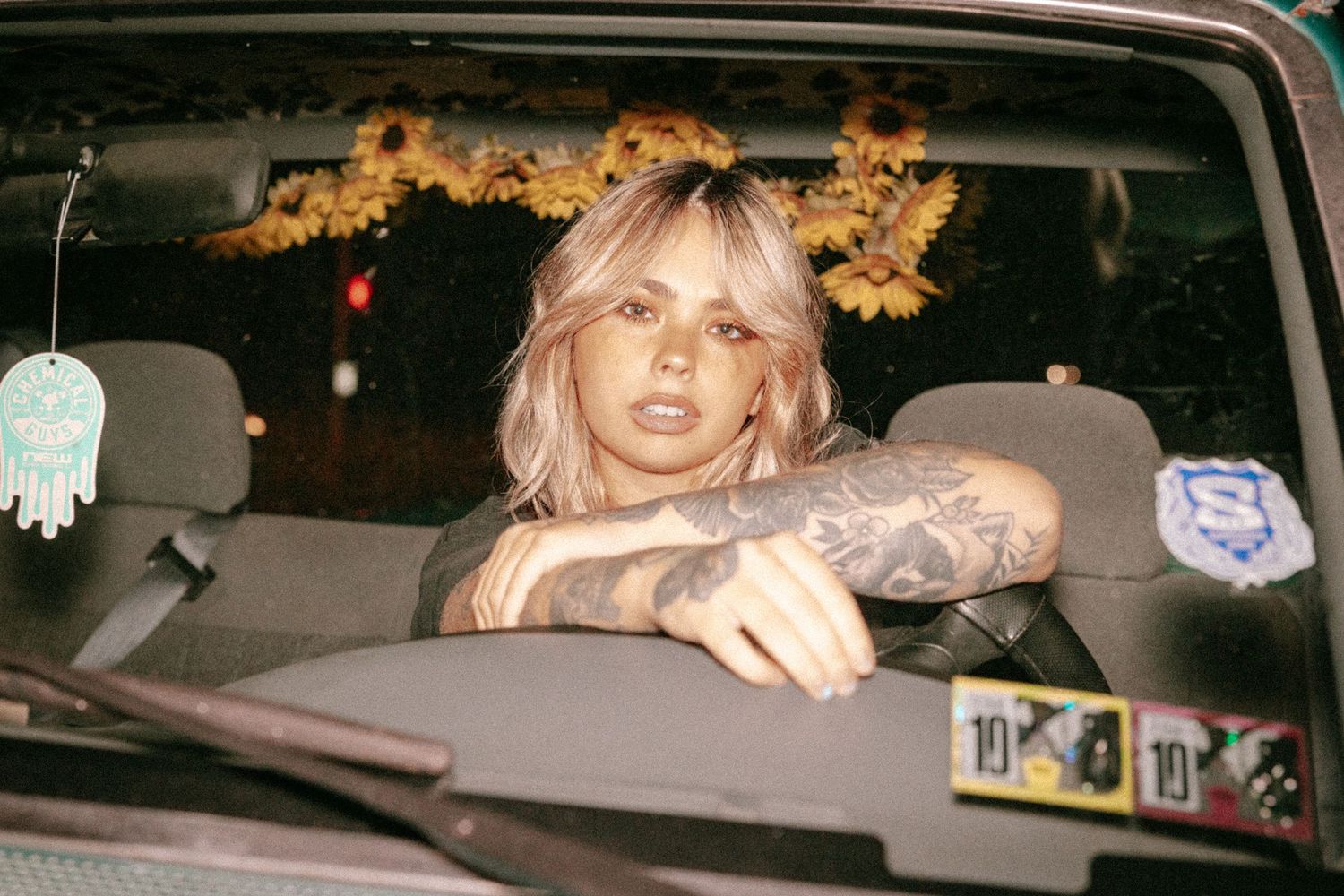 Tattooed blonde woman sitting in car driver seat with sunflower decorations in background, moody aesthetic portrait.