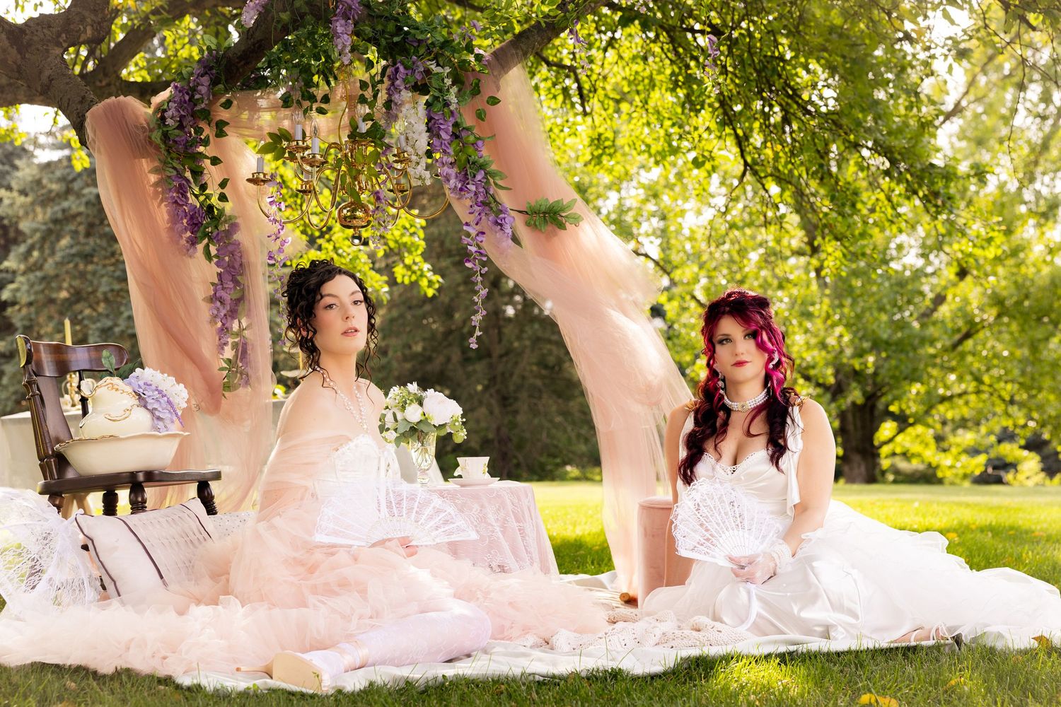 Two brides in white gowns sit on a blanket outdoors under a flowering tree at a garden bridal shoot.