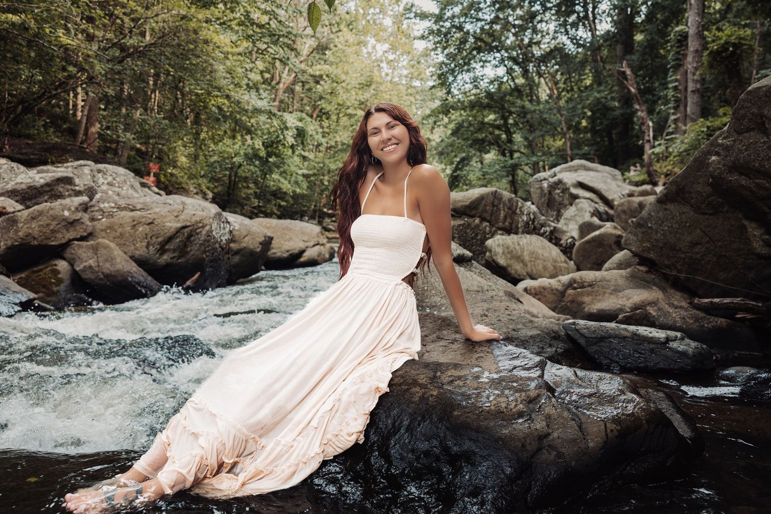 Woman in white dress sitting on rocks by a flowing river surrounded by lush green forest.