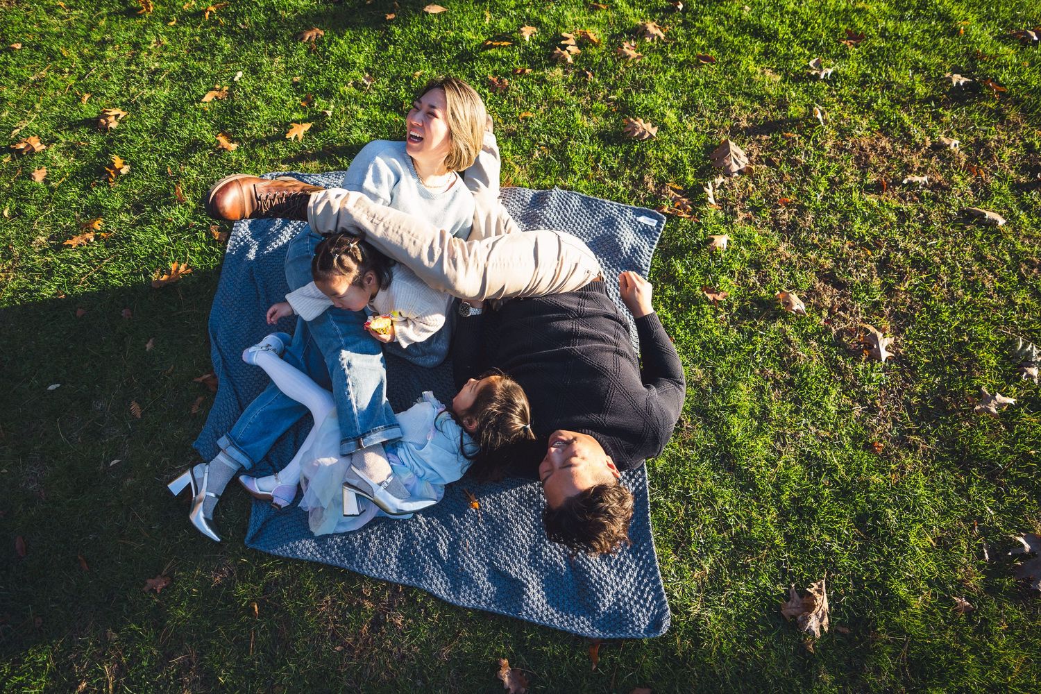 Family relaxing on picnic blanket in grass with fallen autumn leaves.