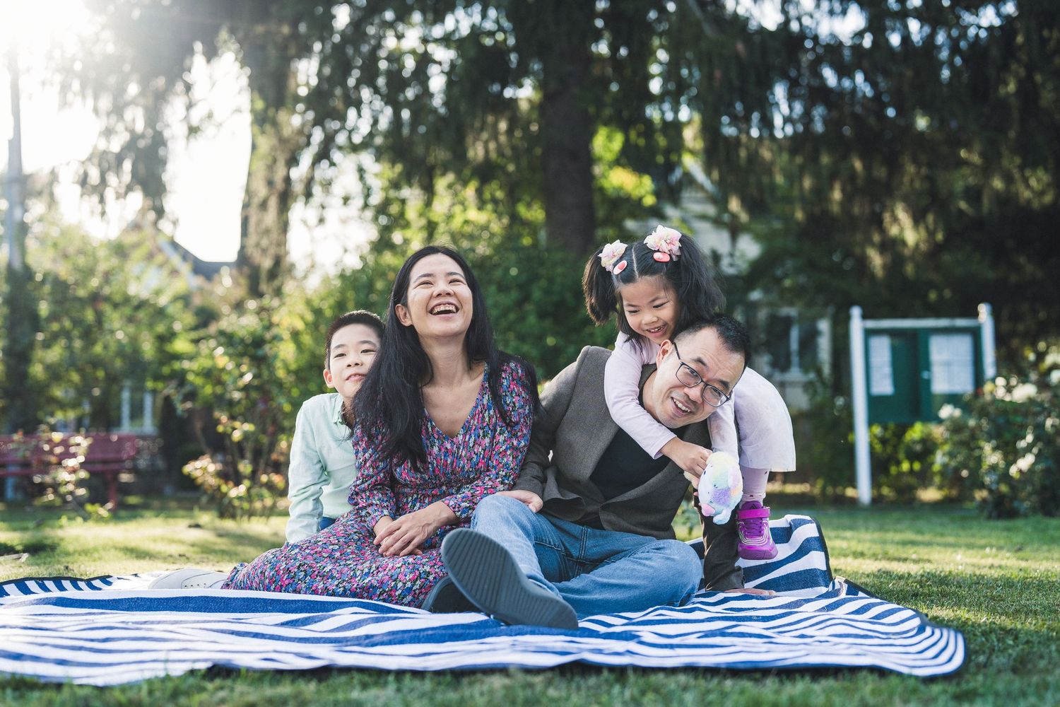 Family picnic scene on striped blanket under tall evergreen trees.