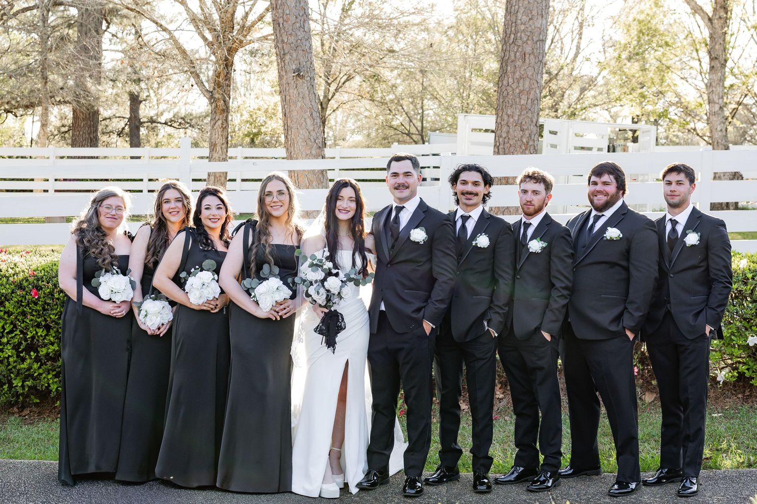 Wedding party posing together in black attire with white floral bouquets at outdoor venue.