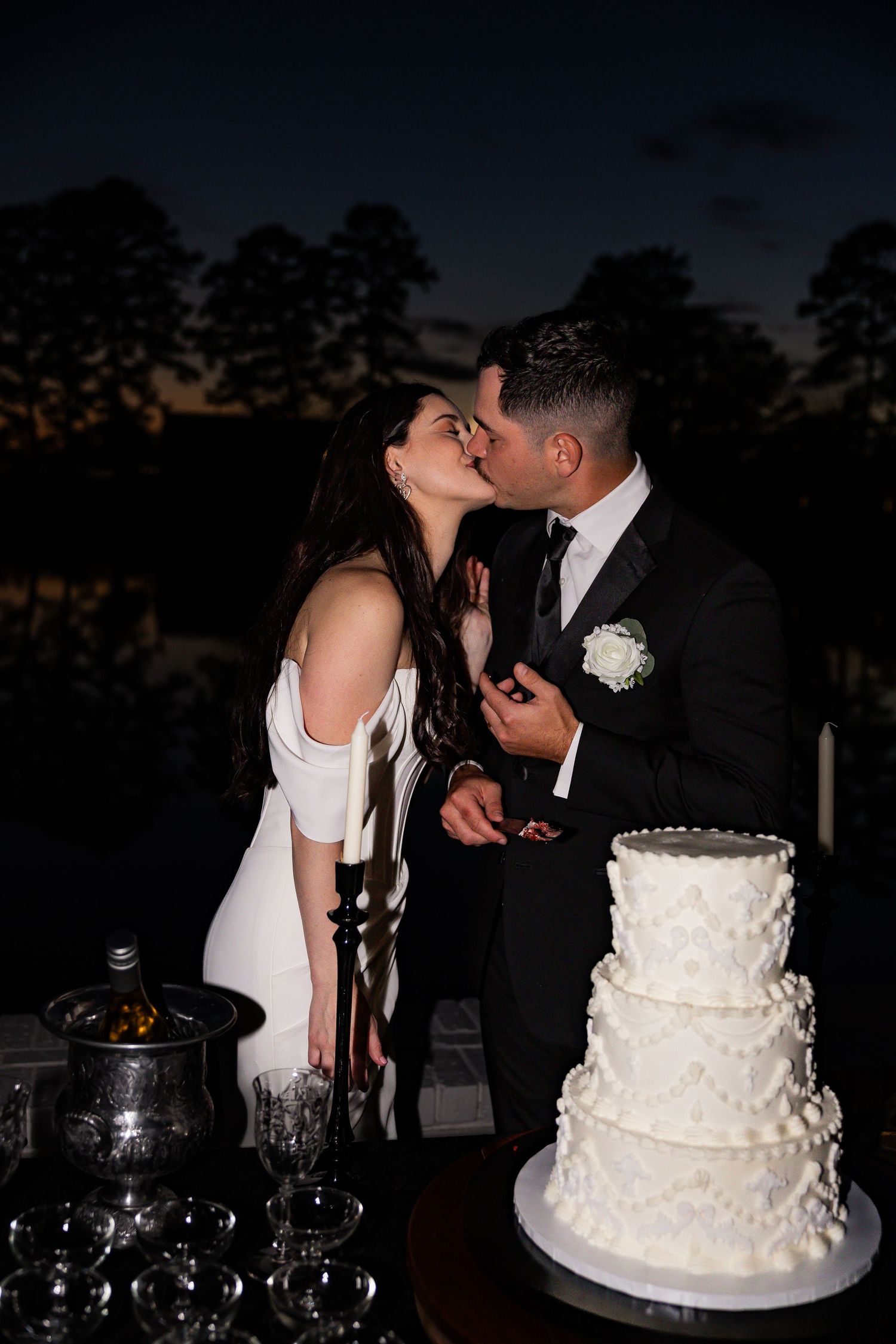 Bride and groom sharing a kiss beside their elegant white tiered wedding cake at dusk.