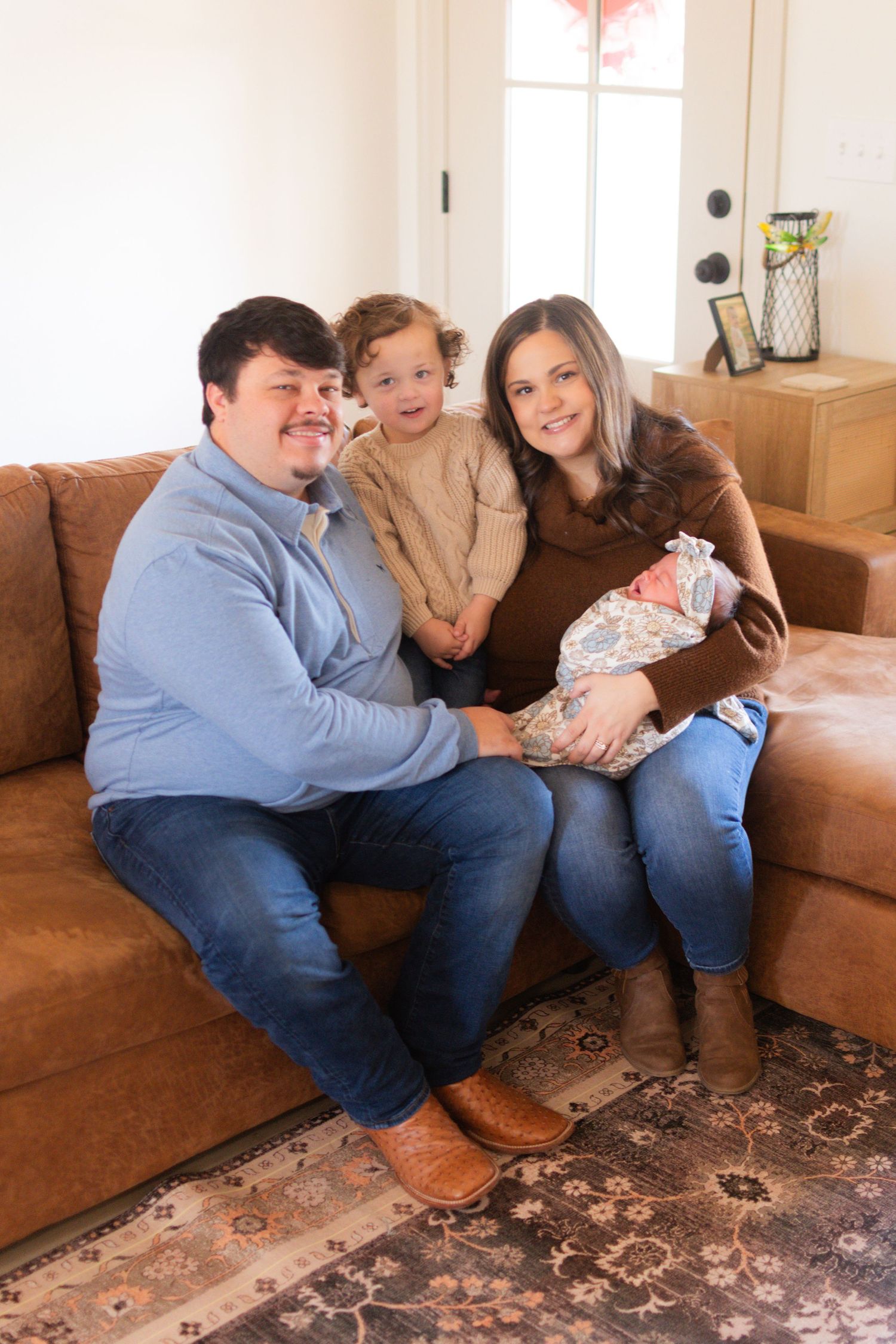 Young family of four sitting on a leather sofa, proudly introducing their newborn baby.