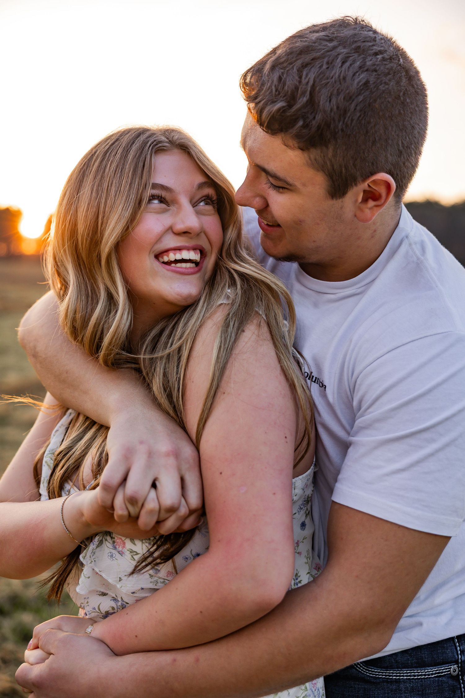Couple laughing and embracing at golden hour in a field, woman in floral dress, man in white t-shirt.