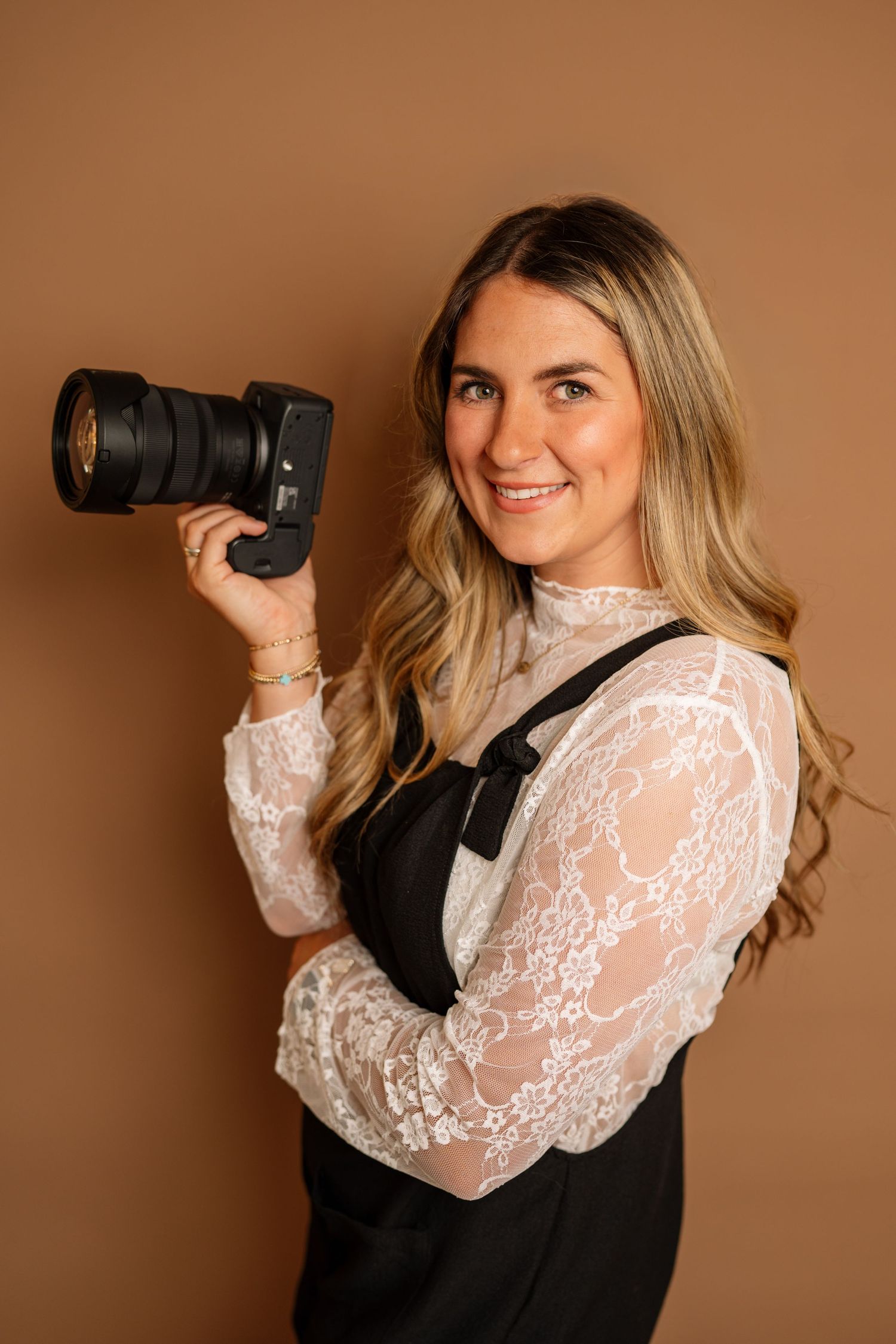 Professional female photographer smiling and holding a DSLR camera against a warm brown background.