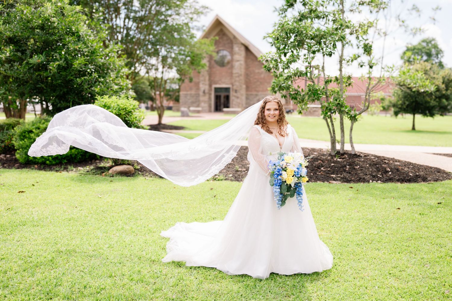 Bride in white gown with flowing veil holding blue bouquet stands on green lawn with stone church backdrop.