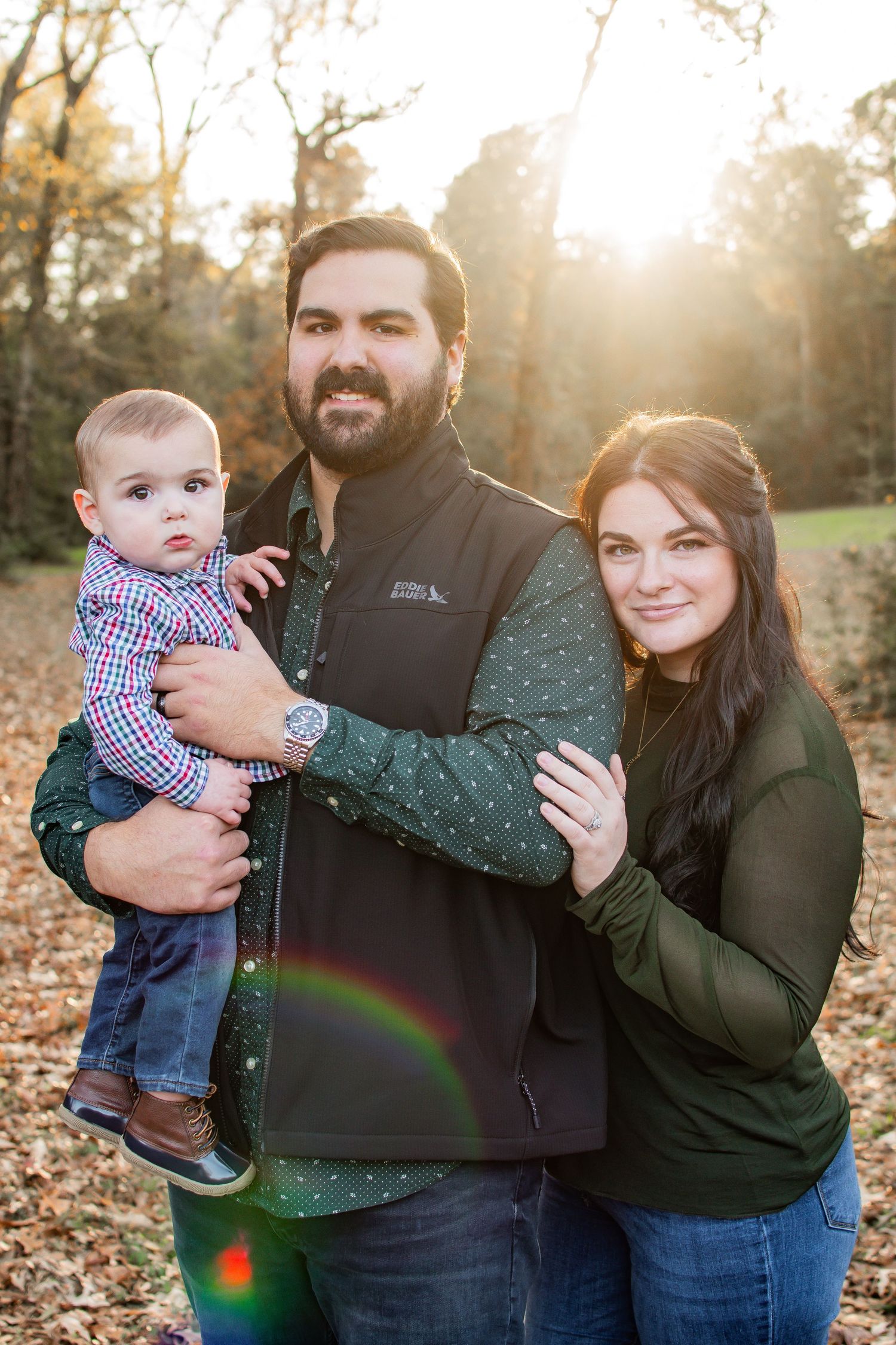Young family portrait with parents holding baby boy outdoors in golden autumn light.
