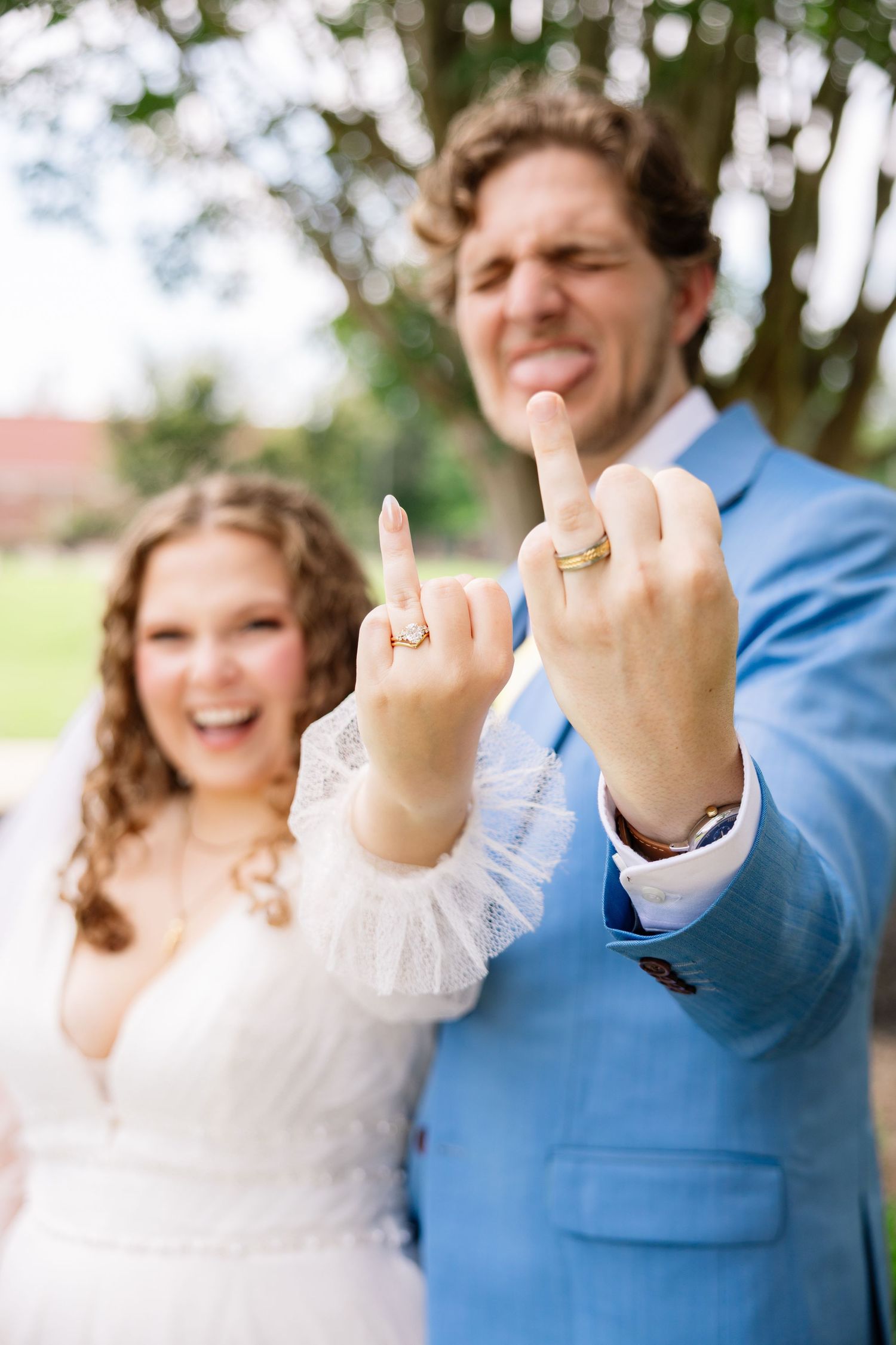 Newlyweds in wedding attire showing off gold wedding rings on their middle fingers, laughing outdoors.