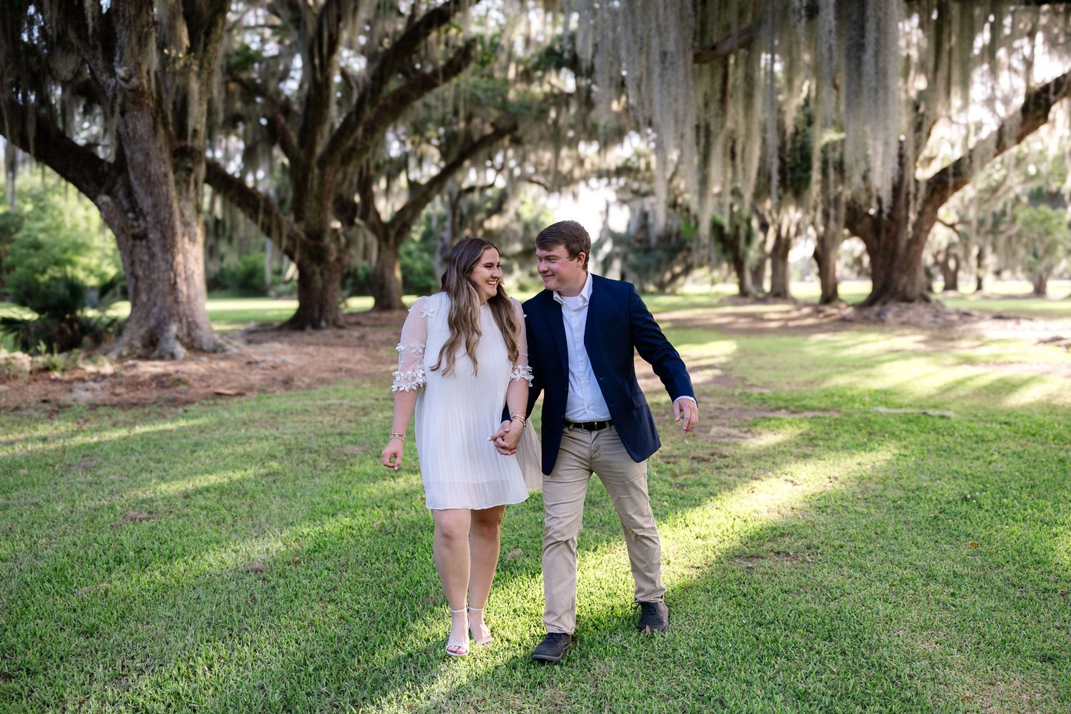 Couple strolling through sunlit park beneath sweeping Spanish moss-draped oak trees.