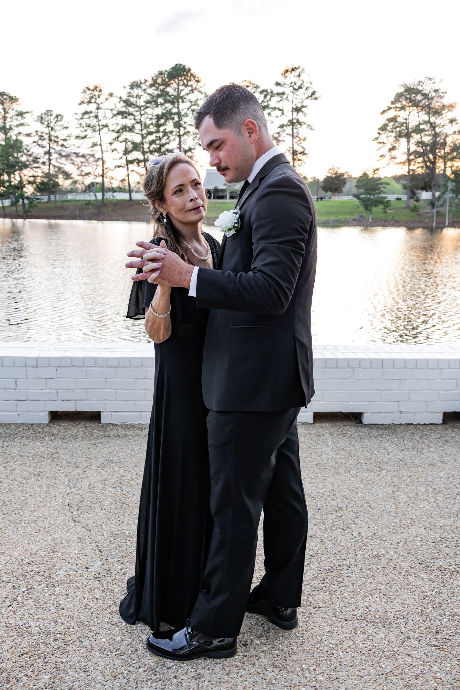 Groom in black tuxedo sharing a tender mother-son dance by a serene lake at sunset.