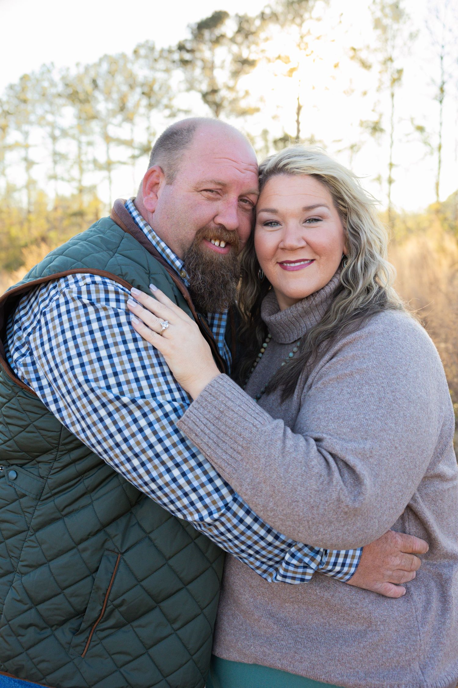 Happy engaged couple embracing outdoors at sunset, woman showing engagement ring while leaning into bearded partner.