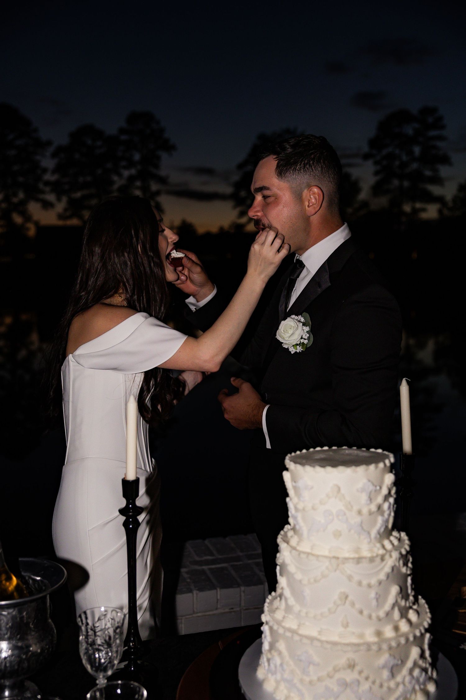 Bride feeding groom a slice of wedding cake beside a white tiered cake at night.