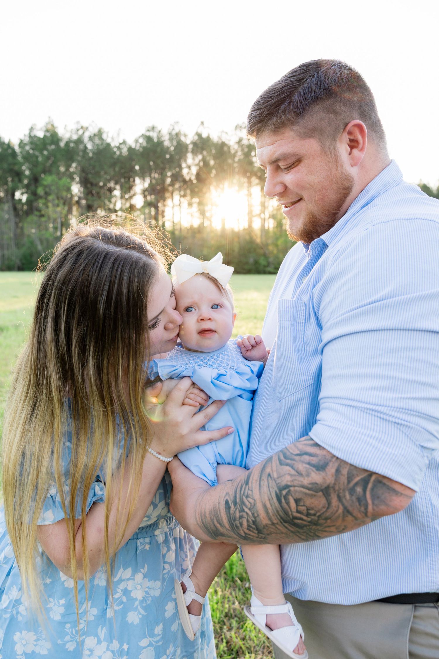 Young family with tattooed father and mother kissing baby girl in blue outfit at sunset.