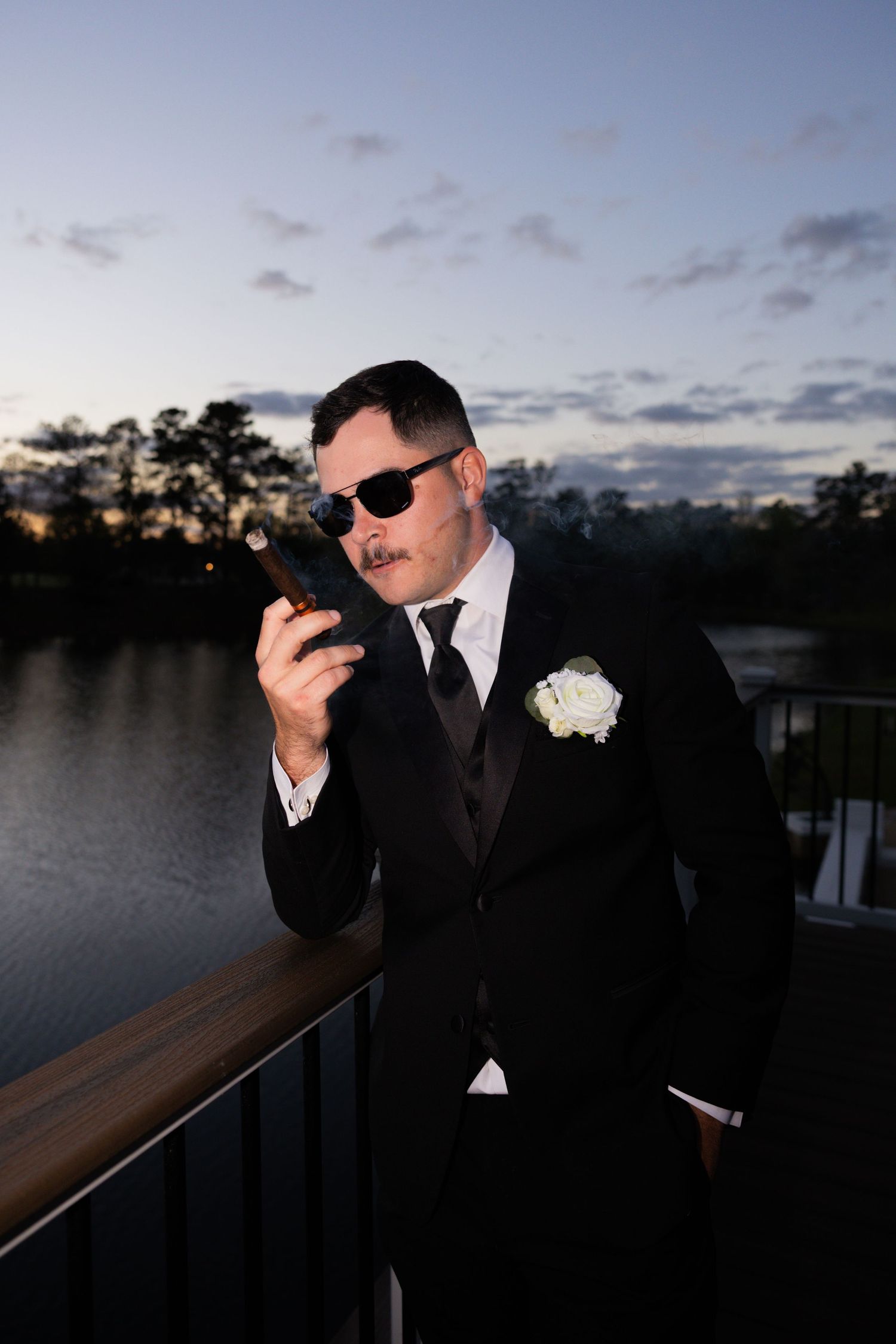 Dapper groom in black tuxedo smoking a cigar on a deck overlooking a lake at dusk.