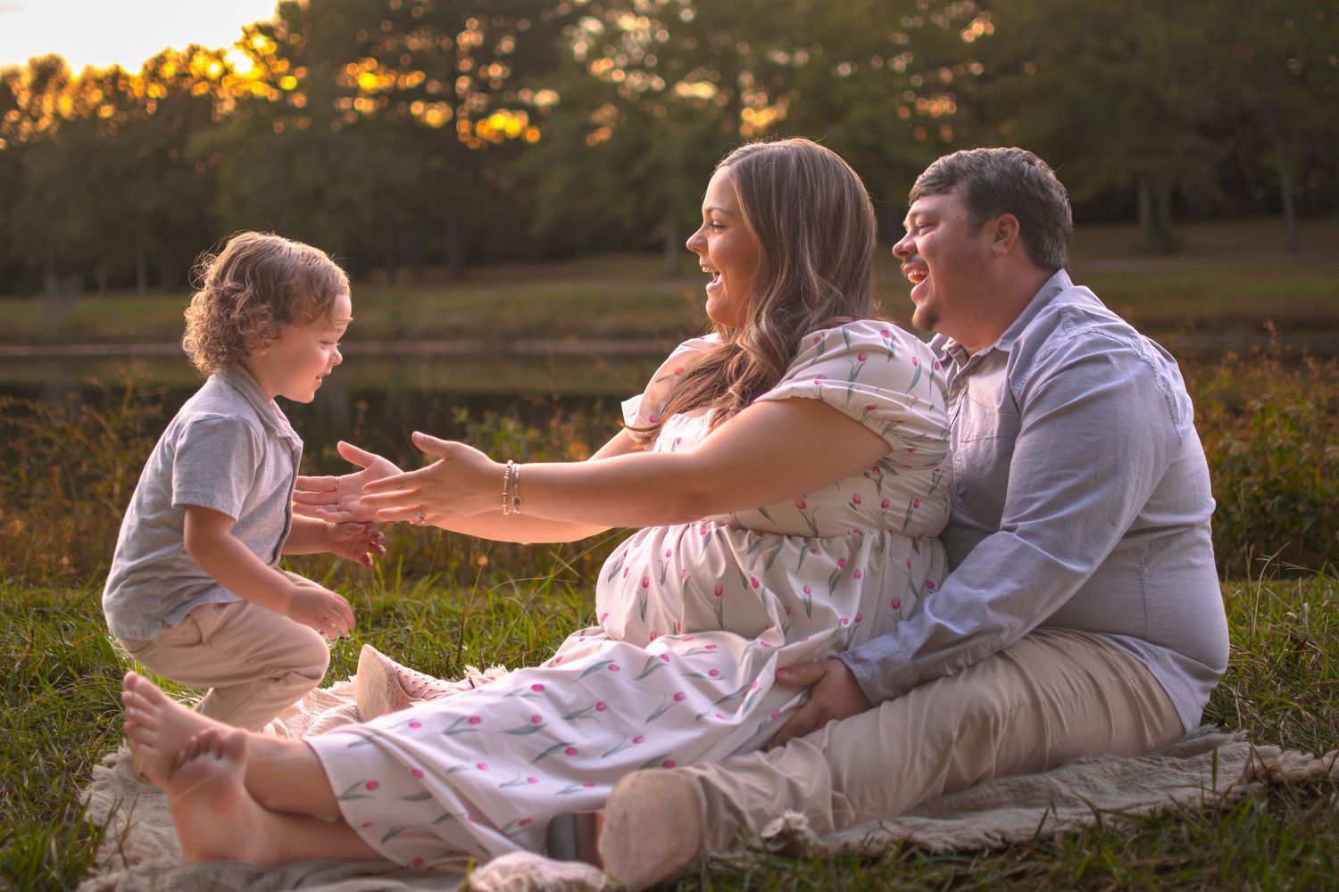 Pregnant woman, husband, and toddler share a joyful moment sitting on blanket in golden field.