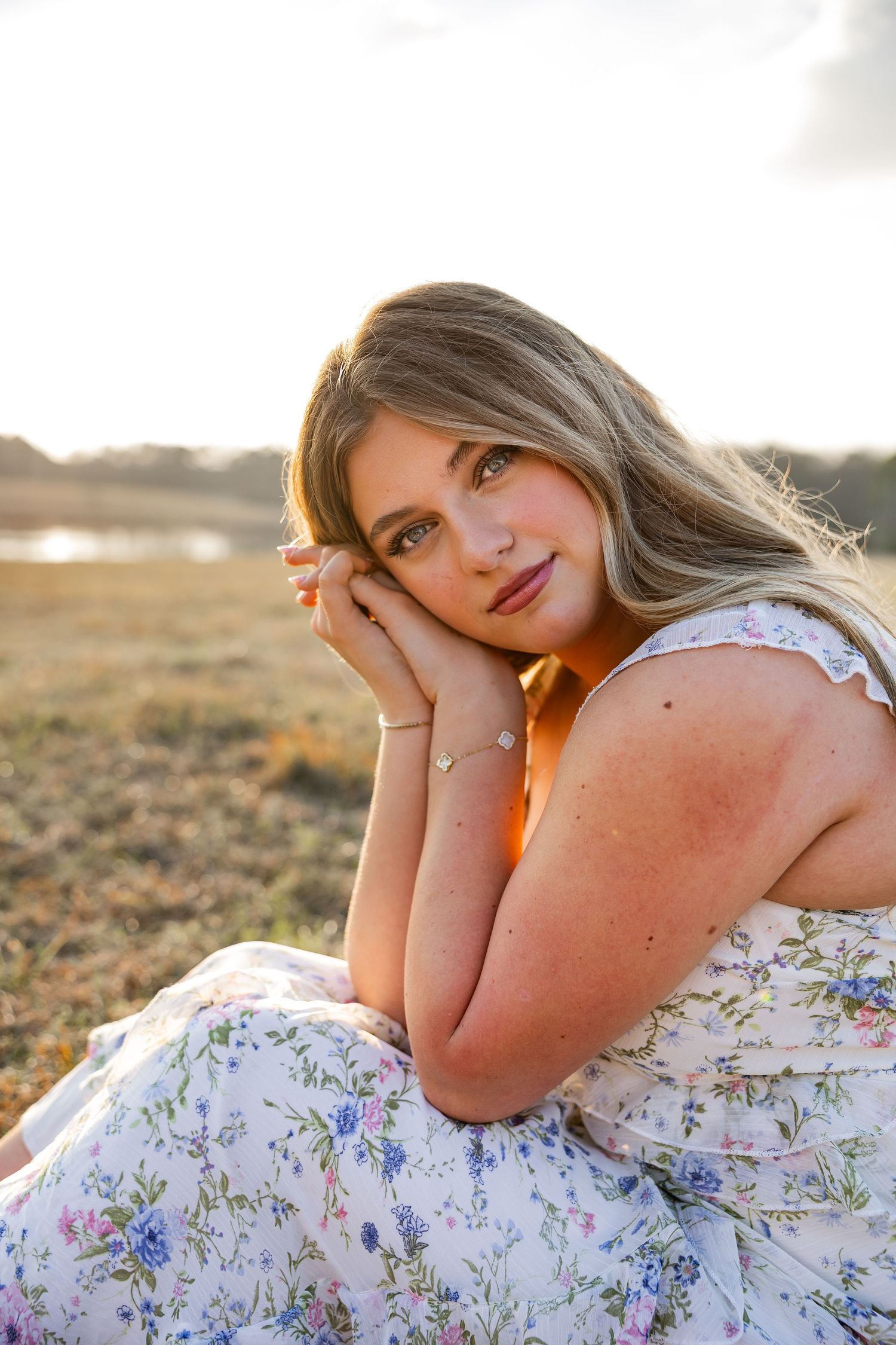 Young blonde woman in floral dress sits in golden field at sunset, resting chin on hands.